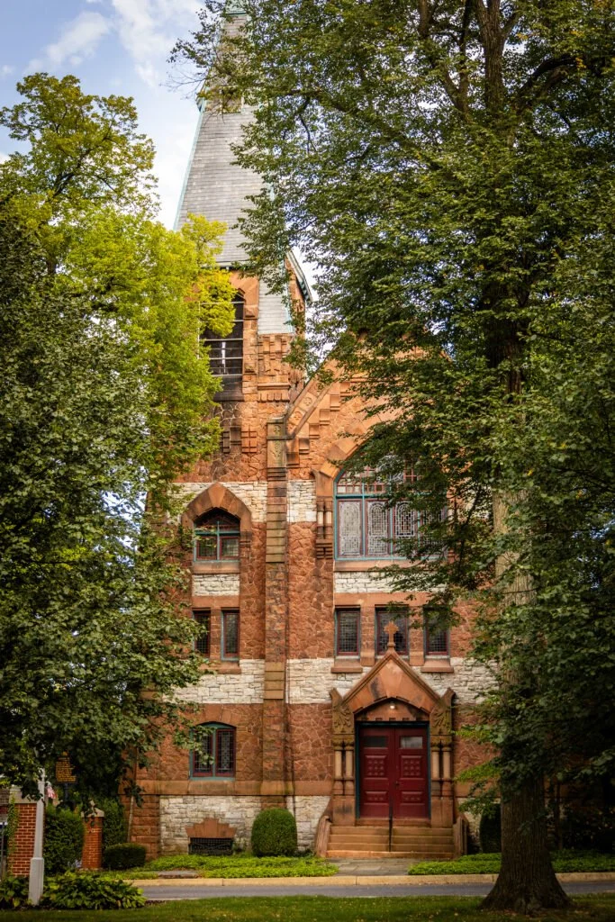 A tall red-brick church with a steep gray roof, surrounded by green trees in a suburban area.