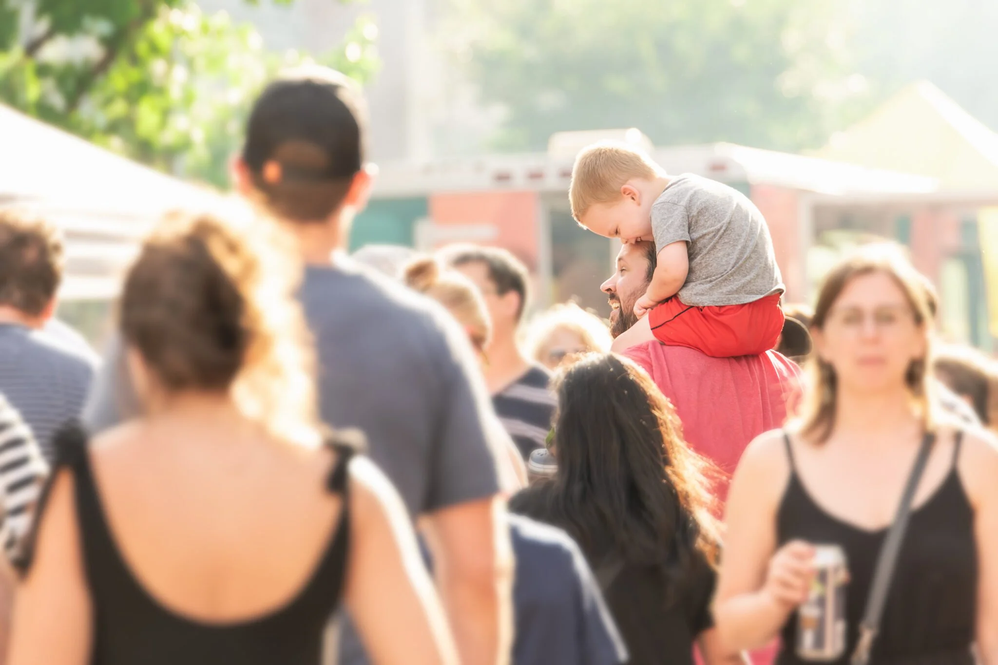 A crowded outdoor event with people talking and socializing, sunlight, and trees in the background. A man is carrying a young boy on his shoulders, and there is a woman holding a can in the foreground.