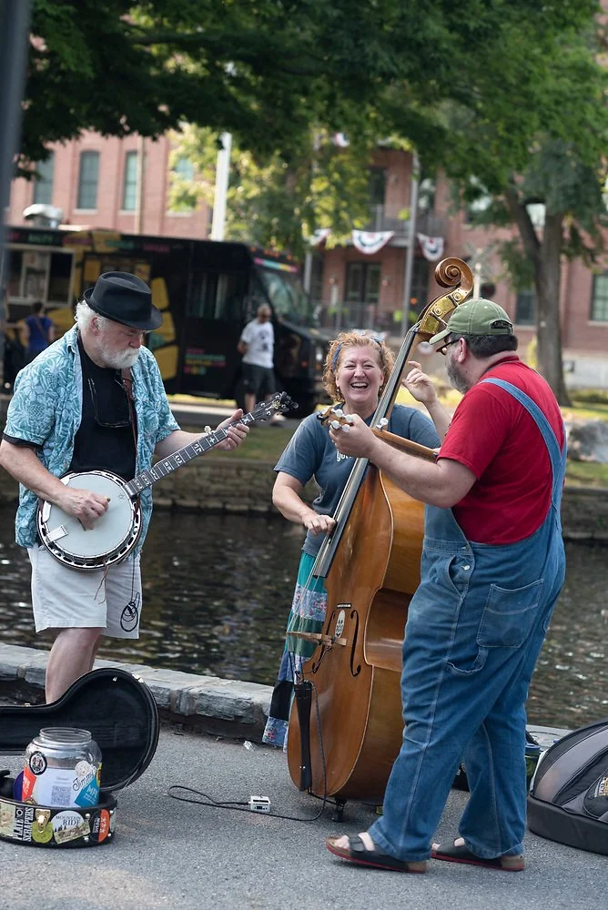 Three street musicians, a man playing a banjo, a woman singing, and a man playing a double bass, perform outdoors near a waterway in a city park, with trees, buildings, and a bus in the background.
