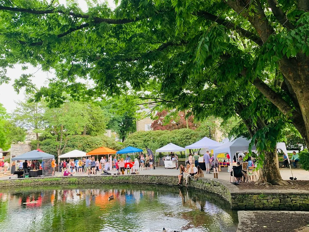 People visiting an outdoor market under colorful tents around a pond in a park with trees.
