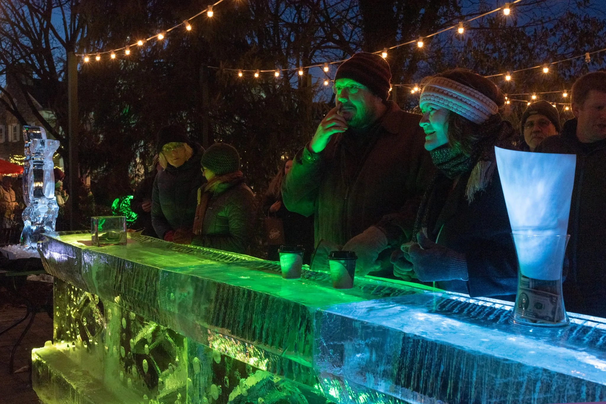 People gathered outdoors at night under string lights, ice bar with illuminated green and blue lighting, and ice sculptures.