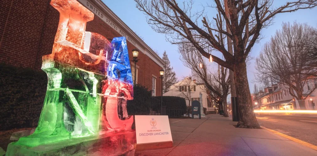 An ice sculpture spelling 'LOVE' with colorful lighting, situated on a sidewalk in front of a brick building and leafless trees at dusk.