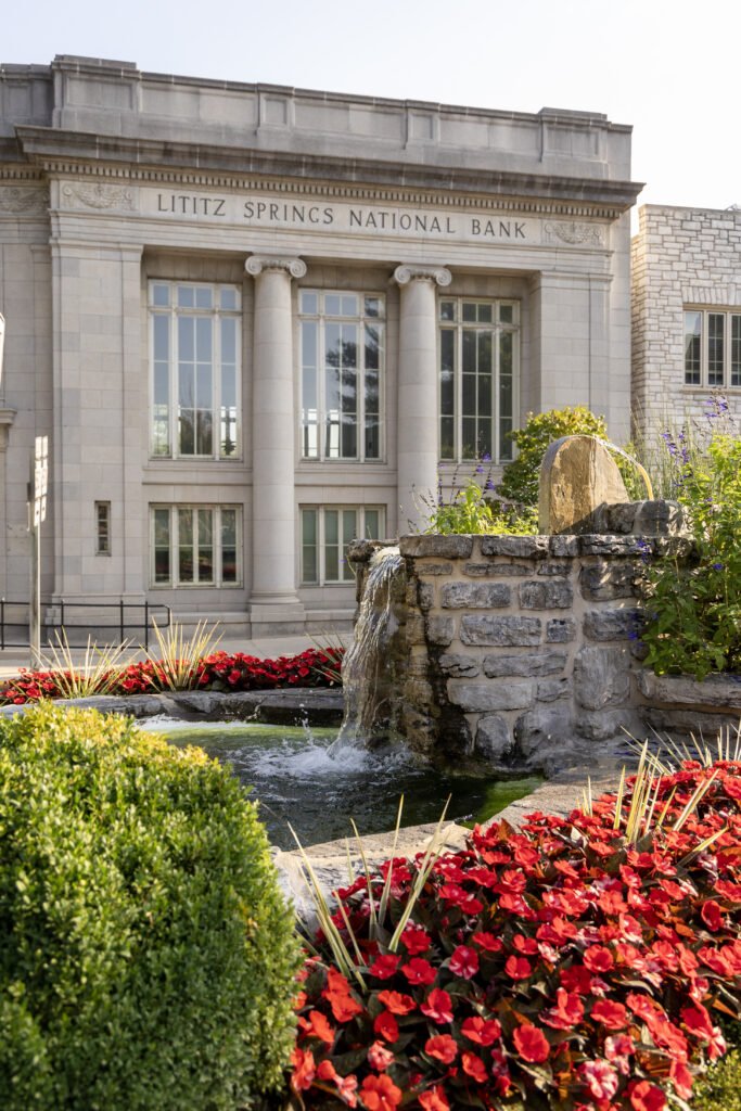 Front view of the Lititz Springs National Bank building with neoclassical architecture, including columns and large windows, surrounded by a landscaped area with red flowers, green bushes, and a stone fountain.