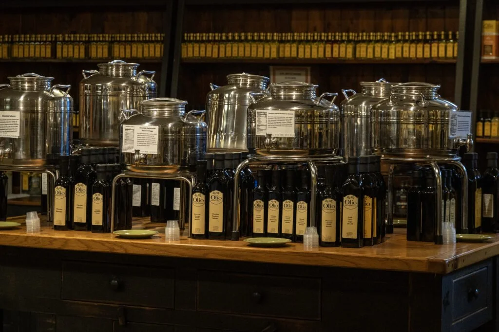 Display of multiple large stainless steel dispensers and bottles of olive oil on a wooden table in a store or cafe setting.