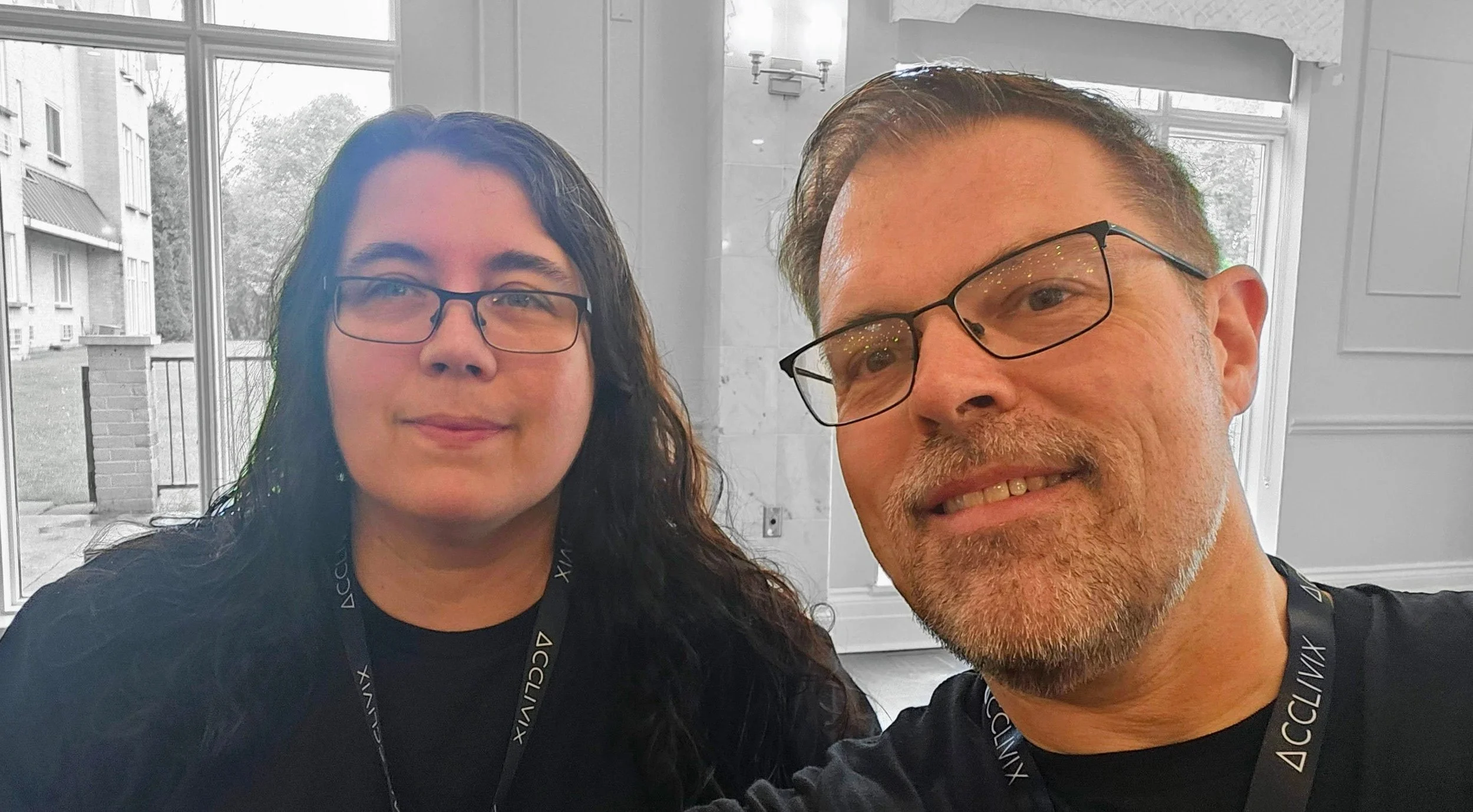 A man and woman taking a selfie indoors with large windows in the background, the woman has long dark hair and glasses, and the man has short hair, glasses, and a beard.