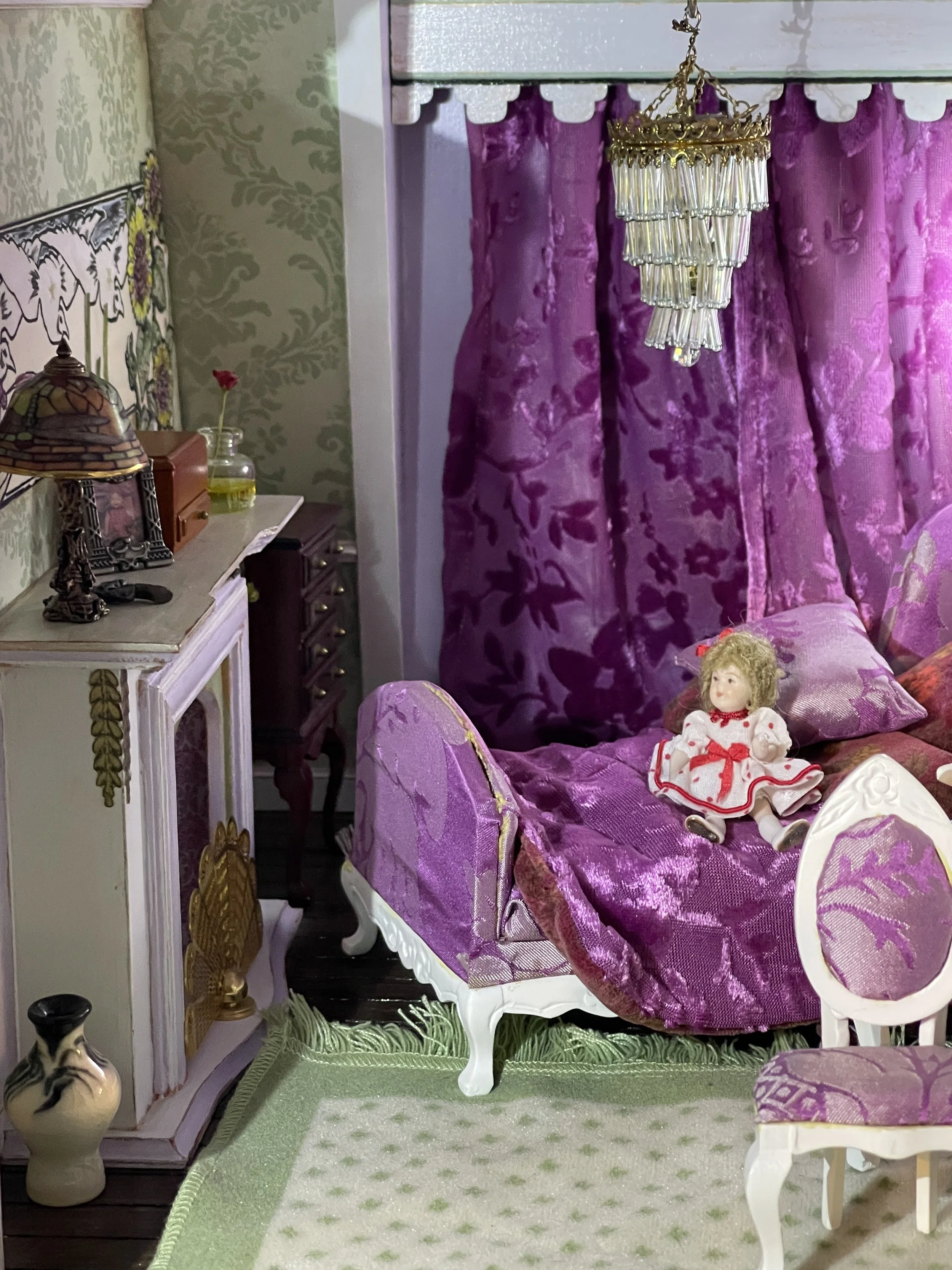 Detail of a bedroom in a miniature dollhouse featuring green brocade walls with matching violet magenta brocade curtains and upholstered bed.  A glass chandelier hangs above the bed with a little doll looking out, designed and painted by Sara Trotter