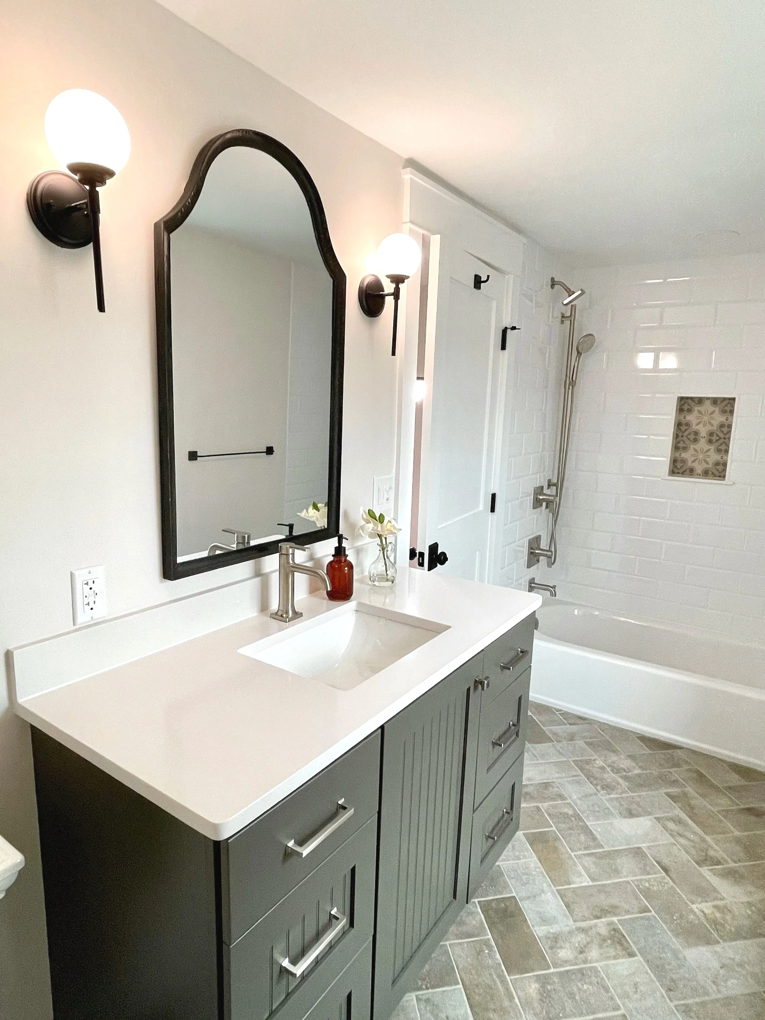 Hall bath with globe pendant lights on either side of a black matte mirror.  The floors are gray brick in a herringbone pattern with beveled white subway tile walls, designed and photographed by Sara Trotter / Chroma Maison Design.