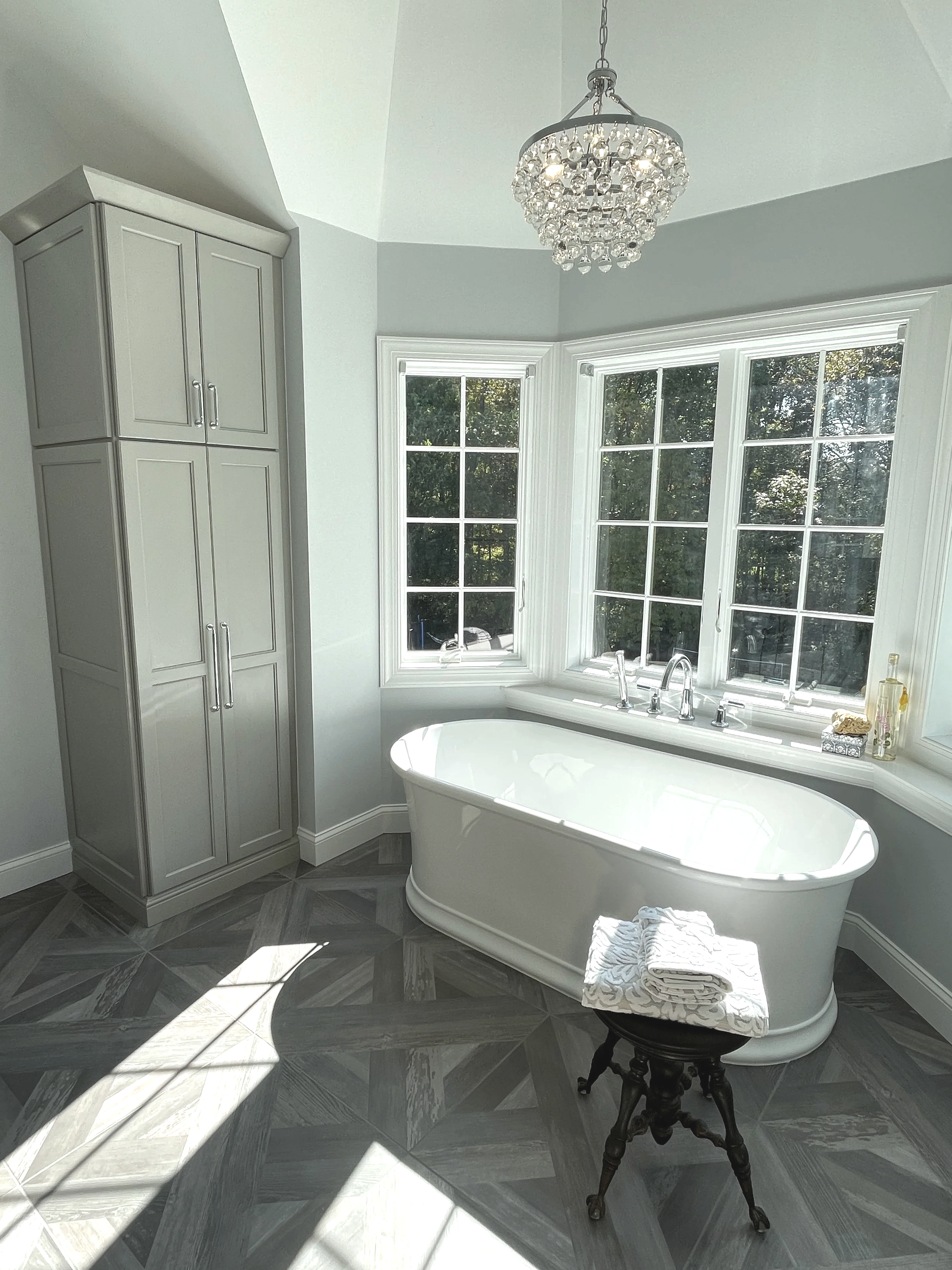 Farmhouse white tub on a distressed blue gray wood parquet tile floor. Above the tub hangs a stunning raindrop crystal chandelier.  What a view out of those windows!  Designed and photographed by Sara Trotter / Chroma Maison Design.