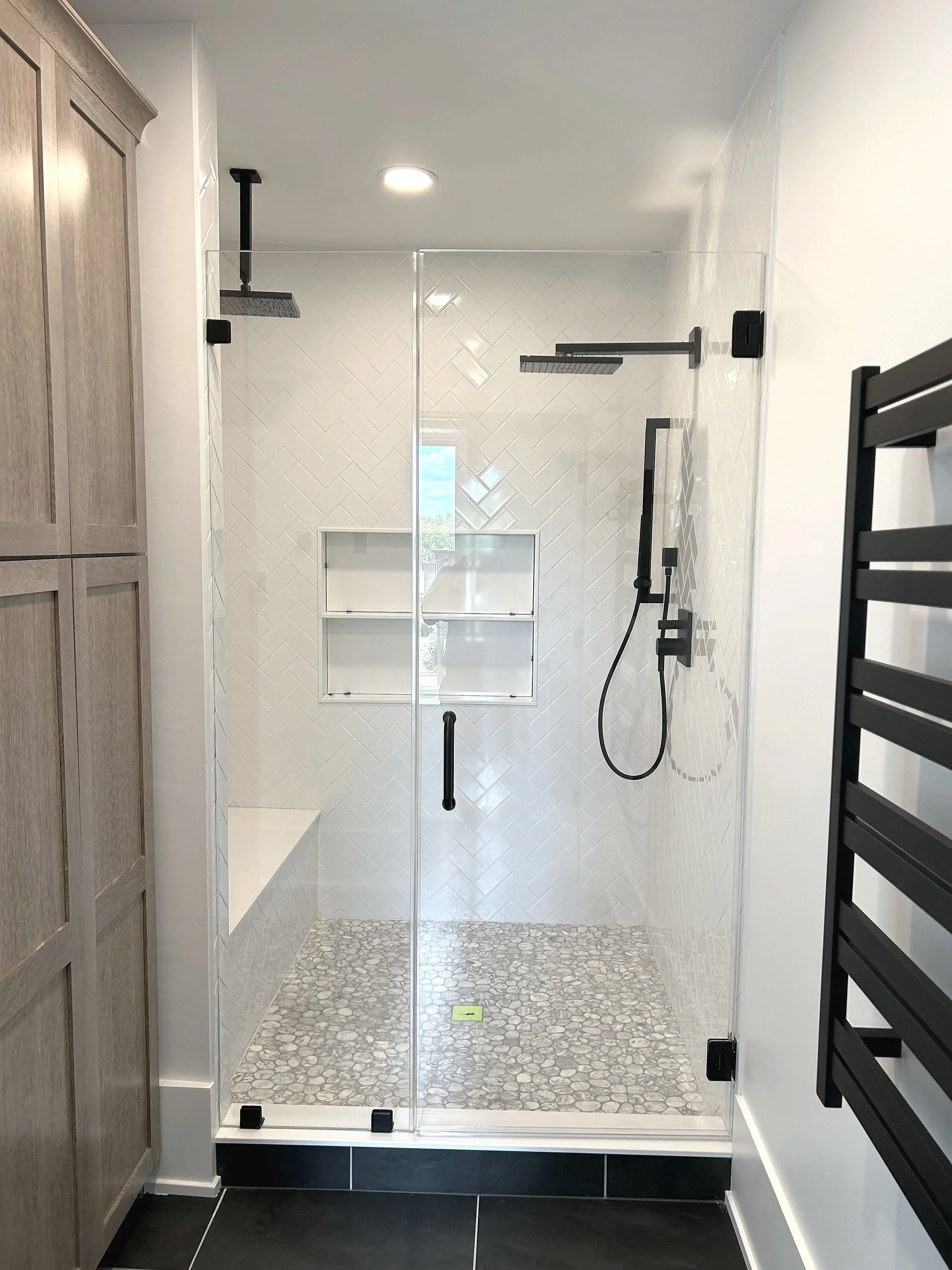 White herringbone pattern subway shower walls with a marble pebble shower floor.  The light shower space is contrasted with the black slate main floor & white oak Omega cabinetry, designed and photographed by Sara Trotter / Chroma Maison Design.