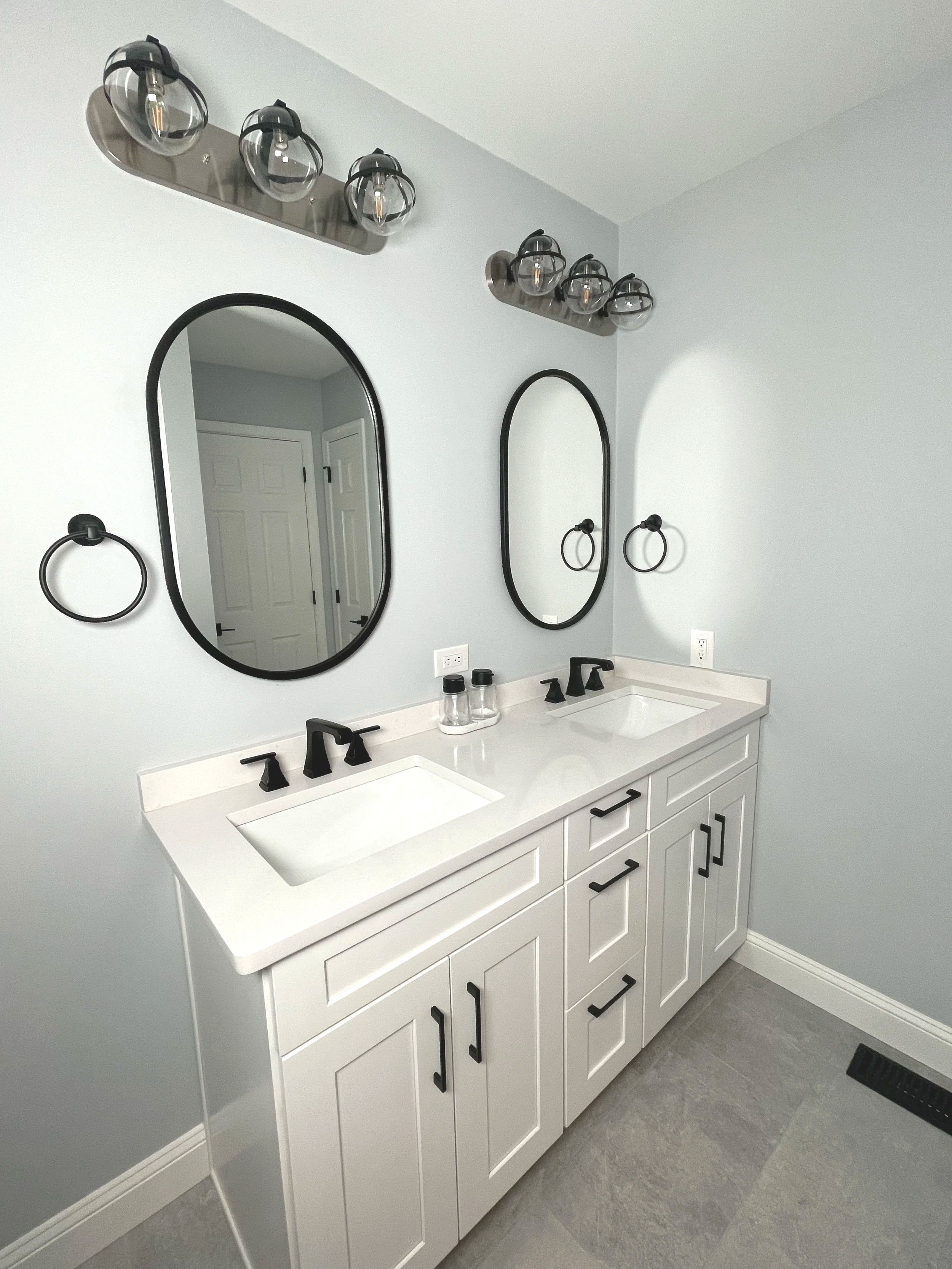 Hall bath with black matte finish clear globe overhead vanity lights above pill shaped mirrors.  The walls are painted in Fleur de Sel, designed and photographed by Sara Trotter / Chroma Maison Design.