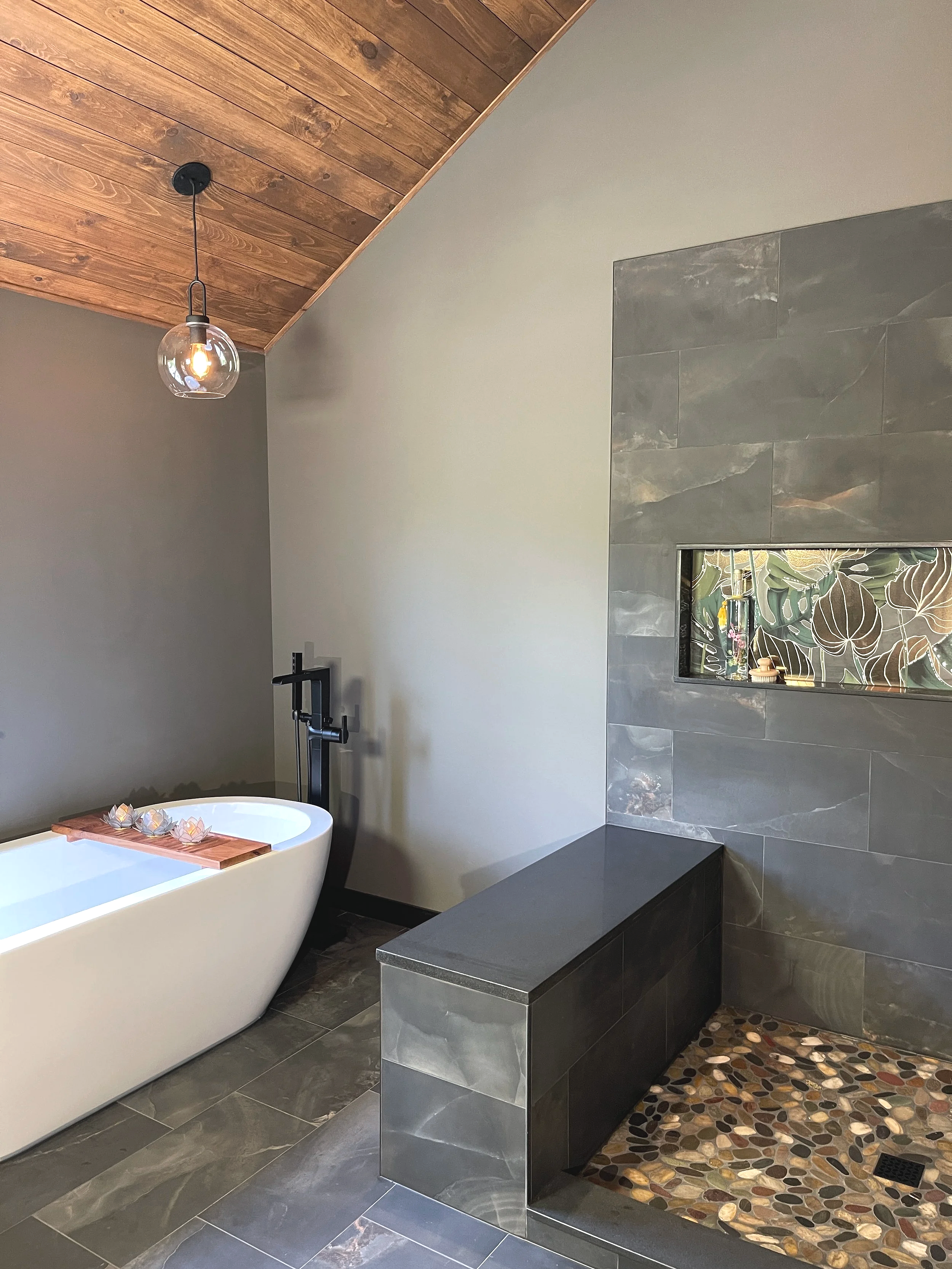 Primary bathroom showcasing black onyx tile shower walls and a colorful pebble floor.  The oversized niche box features embossed Tropez Foliage Oro Artisan tile.  This moody spa-like space has warm gray green walls and a wood ceiling, designed and ph