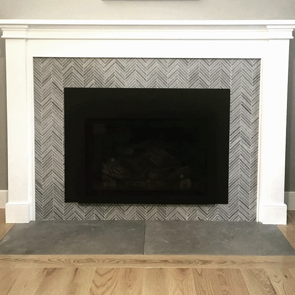 Herringbone pattern gray marble tile around a black fireplace insert with a white mantle, designed by Sara Trotter / Chroma Maison Design.