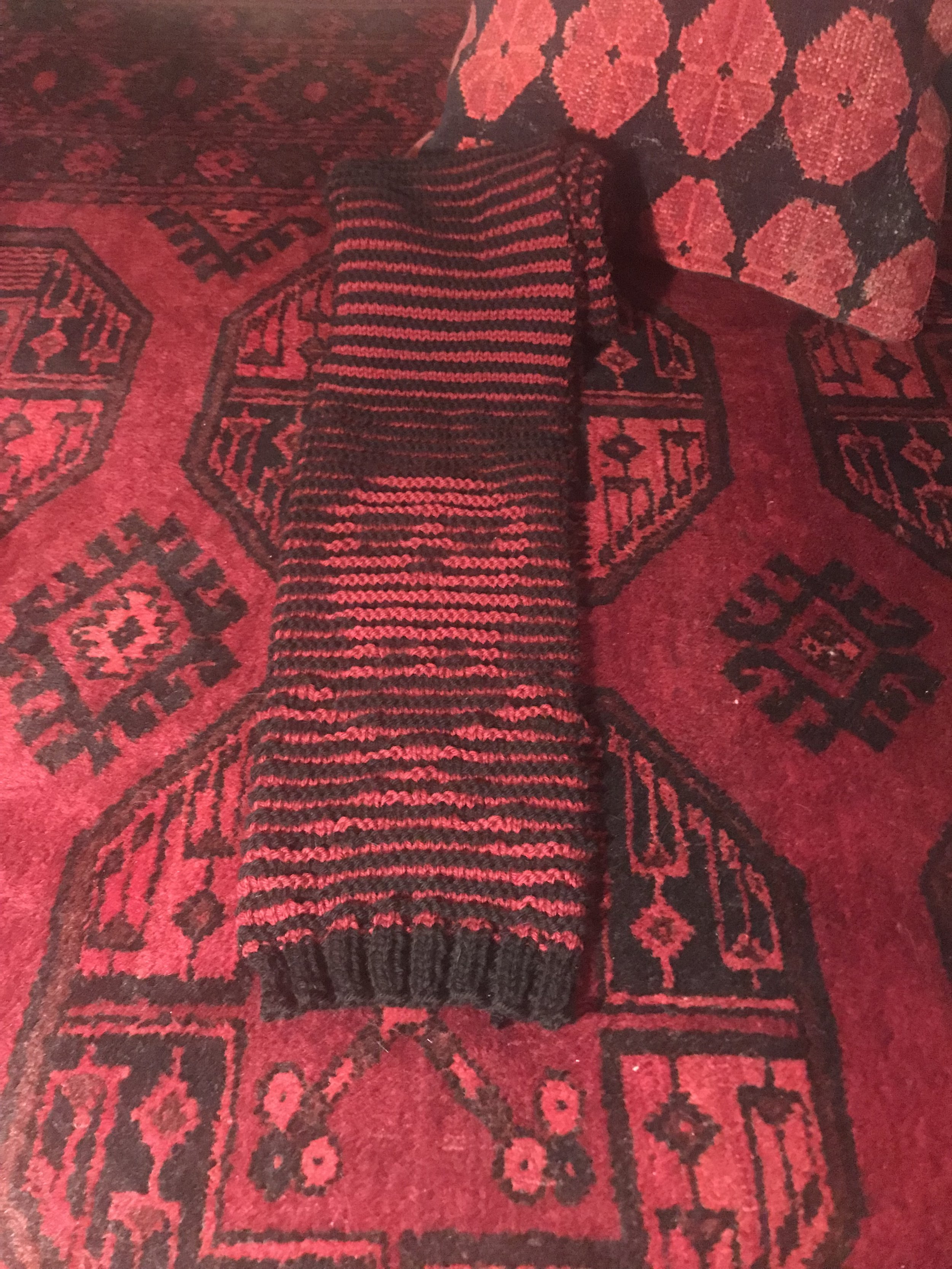 Red and black hand knit illusion skull and cross bones winter scarf displayed on a matching red and black Turkish rug and wool pillow, designed and made by Sara Trotter.