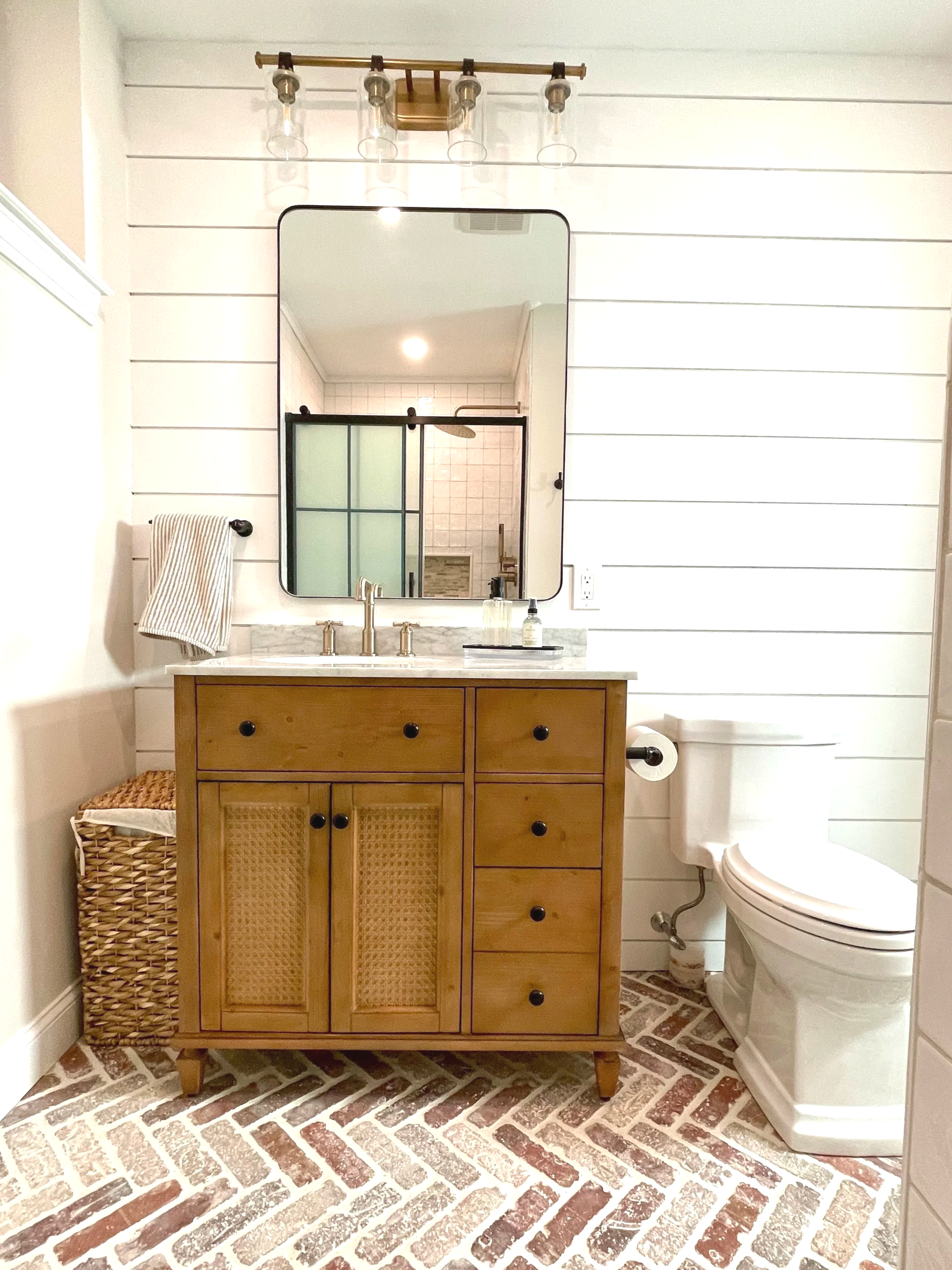 Farmhouse bathroom with a red brick herringbone floor, walnut vanity with caning, and a brass light fixture with leather straps, designed and photographed by Sara Trotter / Chroma Maison Design.