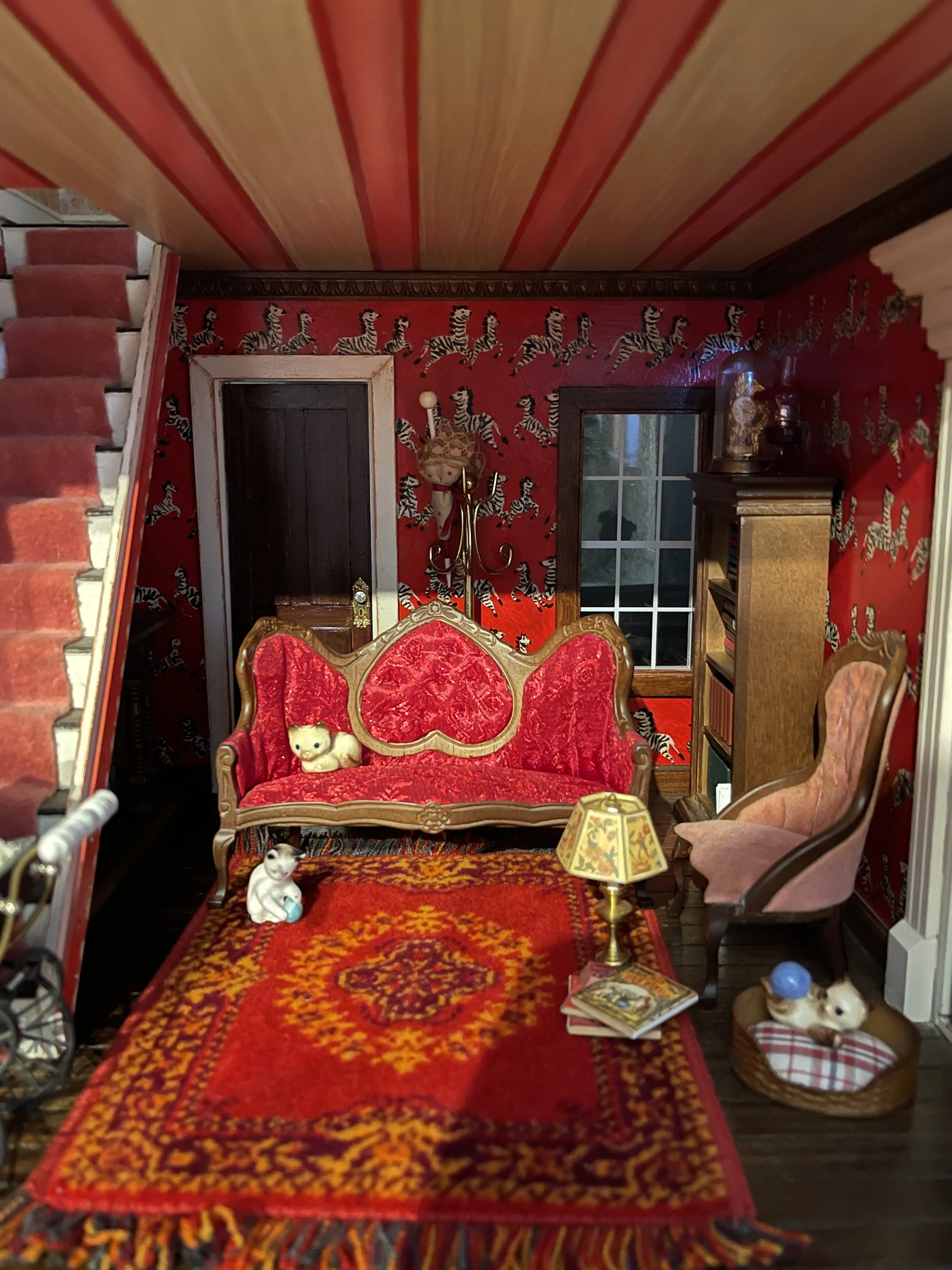 Interior view of a miniature dollhouse room with hand-painted replica Scalamandre Masai Red Zebra wallpaper design.  The ceiling is painted in ribbon stripes with a red heart Victorian love seat and cats playing on a red Oriental area rug, designed a