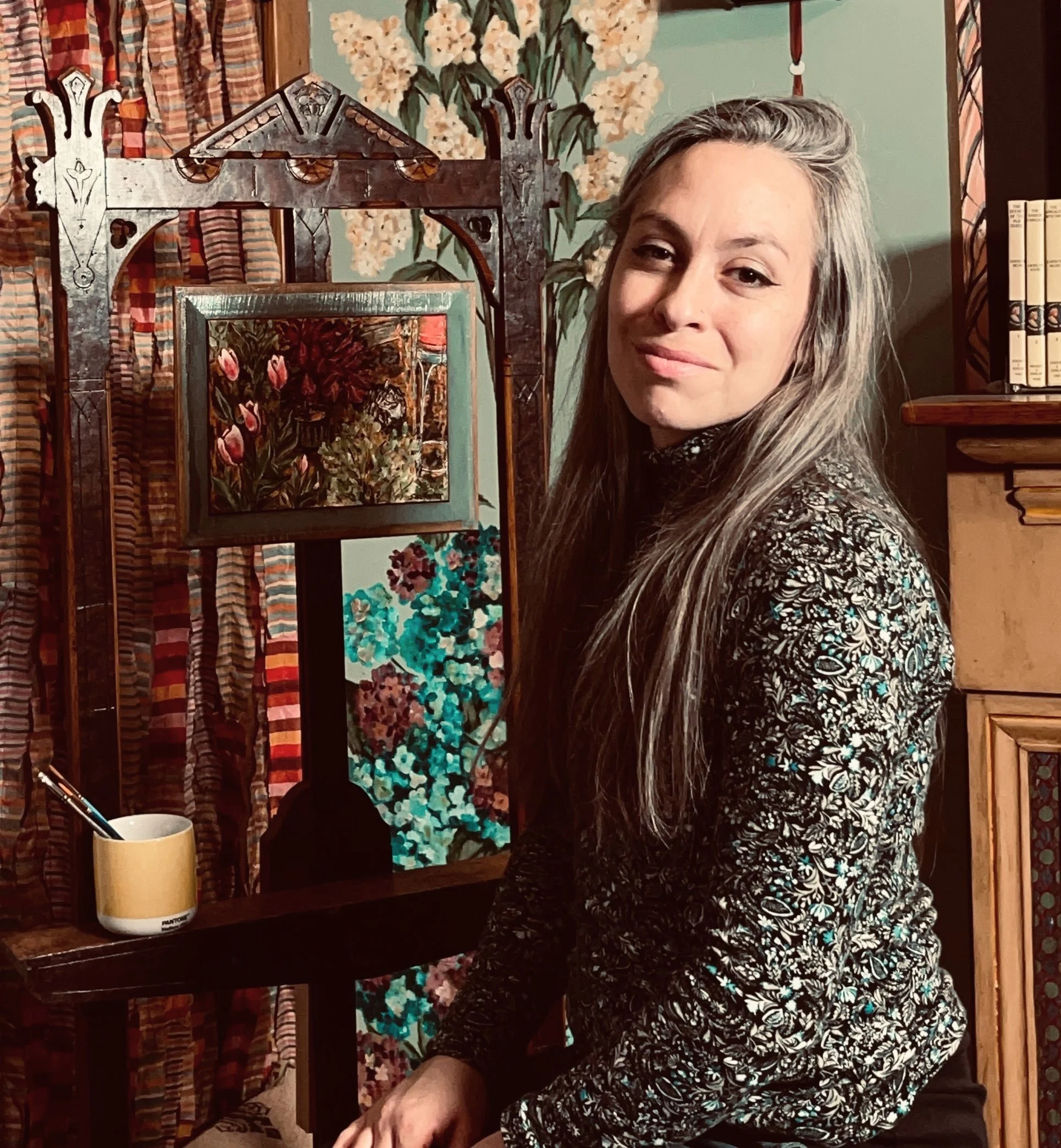 A professional portrait of Sara Trotter, owner of Chroma Maison Design.  She is a young woman with long silvery gray hair sitting beside an Eastlake antique easel with a small painting.