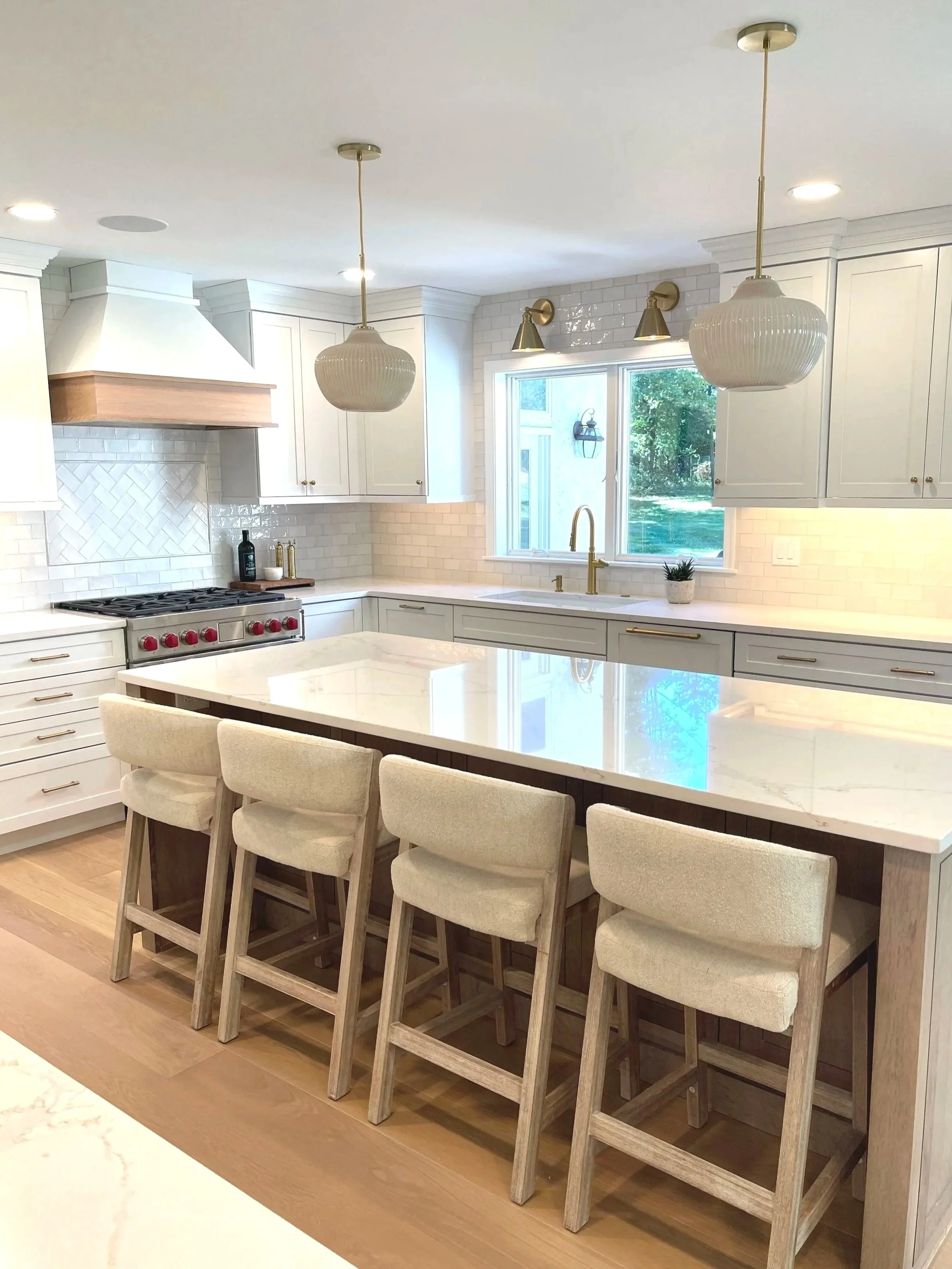Boucle kitchen stools around an Omega white oak island.  Plenty of light with above island pendants and brass sconces by the picture window, designed and photographed by Sara Trotter / Chroma Maison Design.