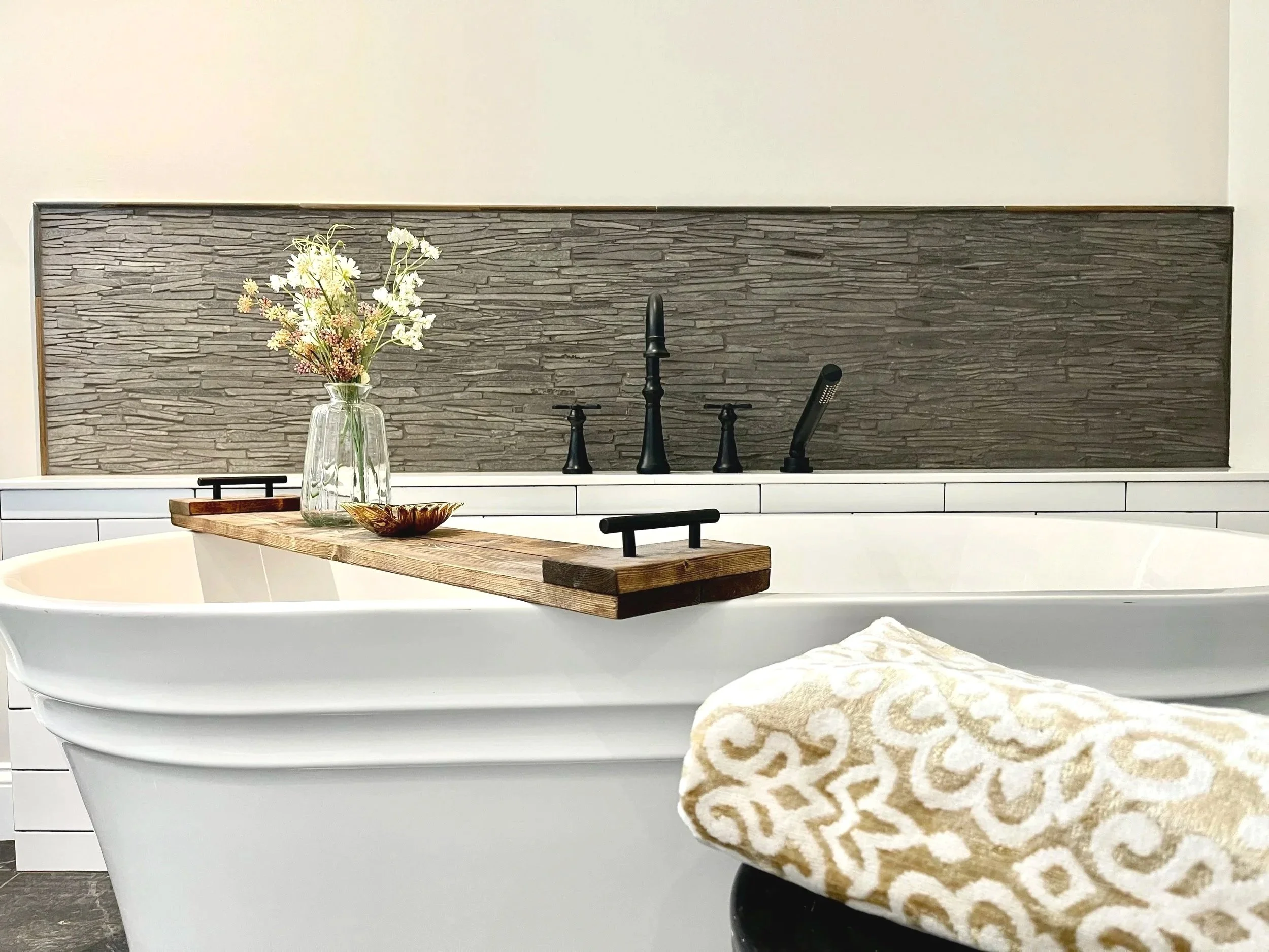 A modern bathroom with a white bathtub, a wooden tray on the tub, a towel with a gold pattern on a black stool, a vase with white flowers, and a dark textured tile backsplash behind a black faucet and fixtures.