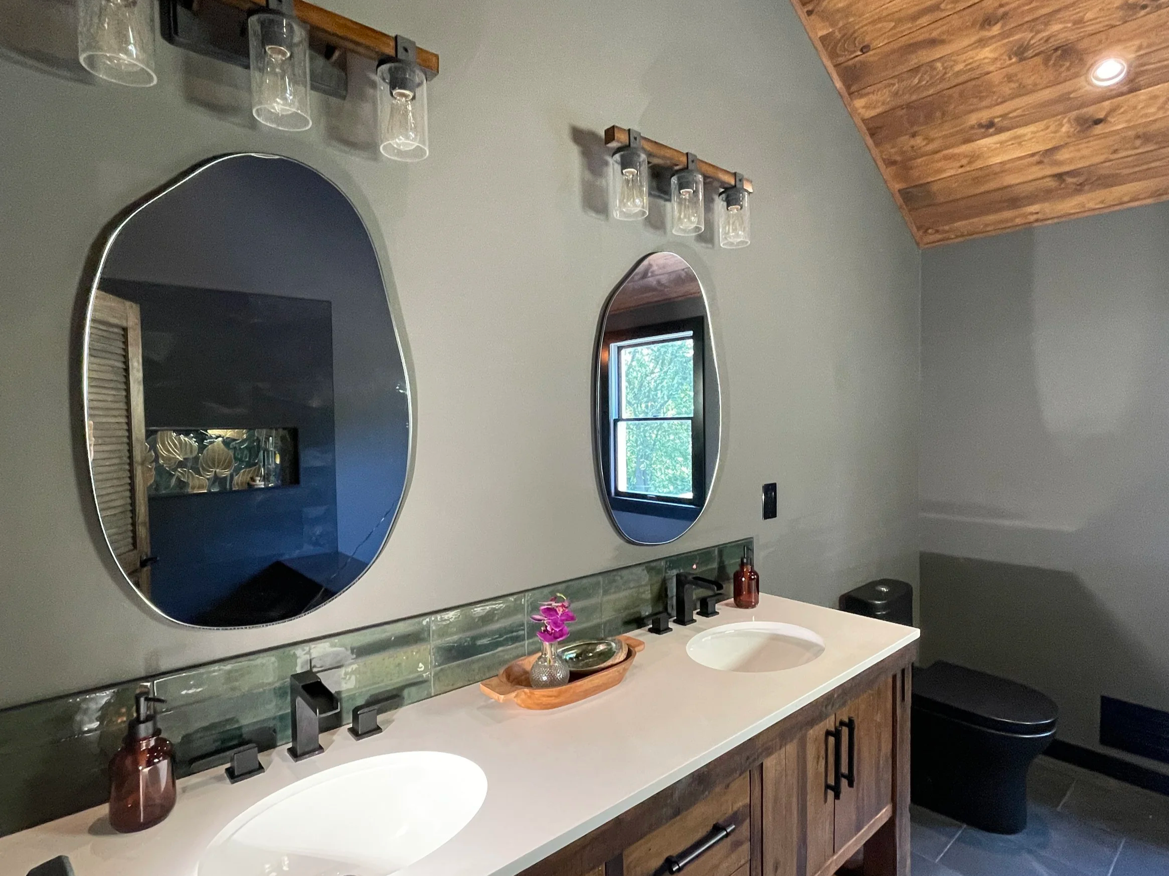Two asymmetrical mirrors above a rustic double vanity featuring a green subway tile backsplash.  In the mirror's reflection we see the large niche box with tile Tropez Foliage Oro Artisan, designed and photographed by Chroma Maison Design.