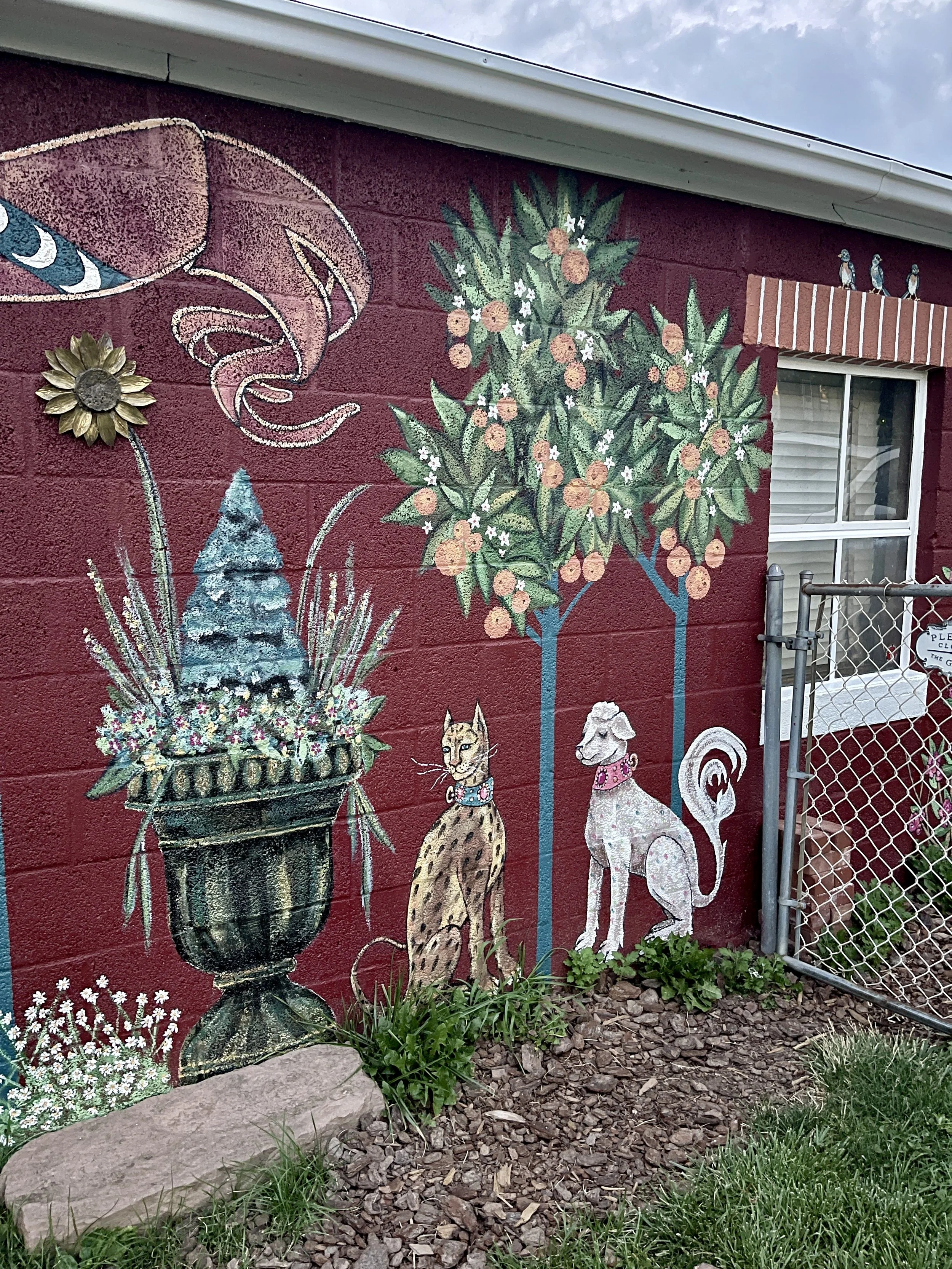 Exterior painted mural inspired by the Unicorn Tapestries featuring an orange tree, a leopard next to a white dog - both with fancy jeweled collars, a moon flag, and three birds against a barn red background.  By artist Sara Trotter / Chroma Maison D
