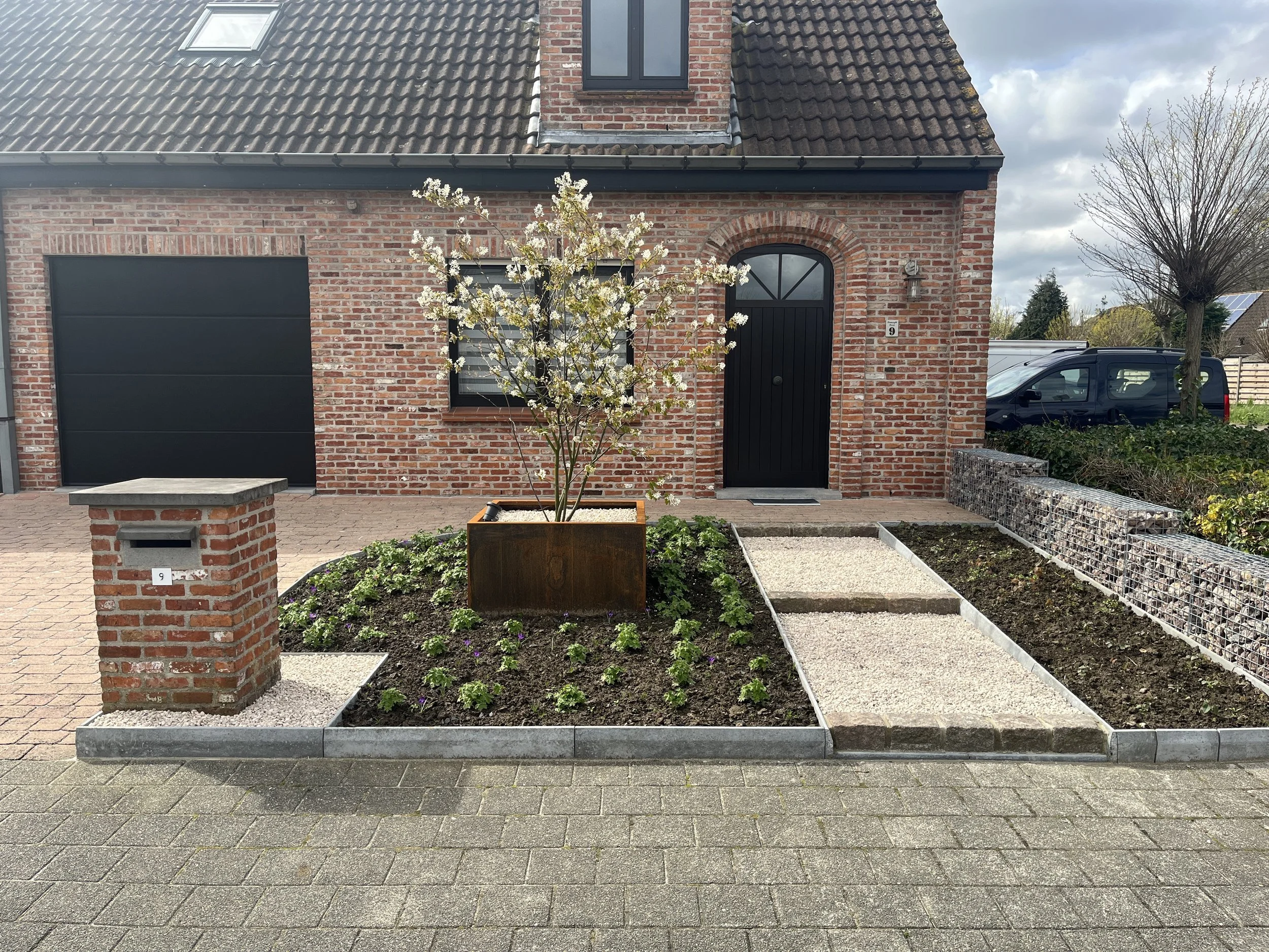 Front yard of a brick house with a black front door, a small flowering tree in a wooden planter, and a garden bed with young plants, gravel pathways, and a gabion wall on the side. A black van is parked in the background.