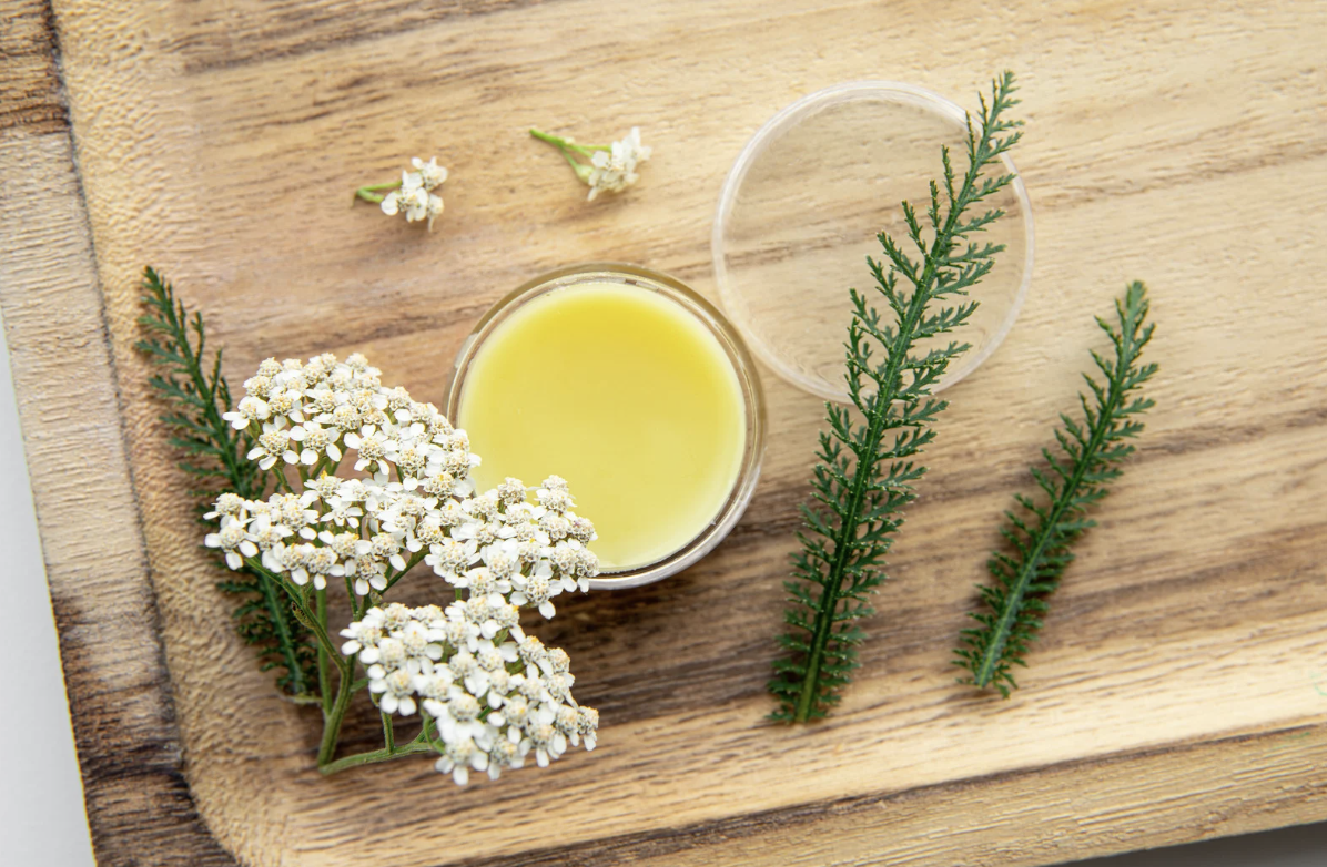 A small open container of yellow herbal oil, surrounded by white flowers and green fern-like leaves on a wooden surface.