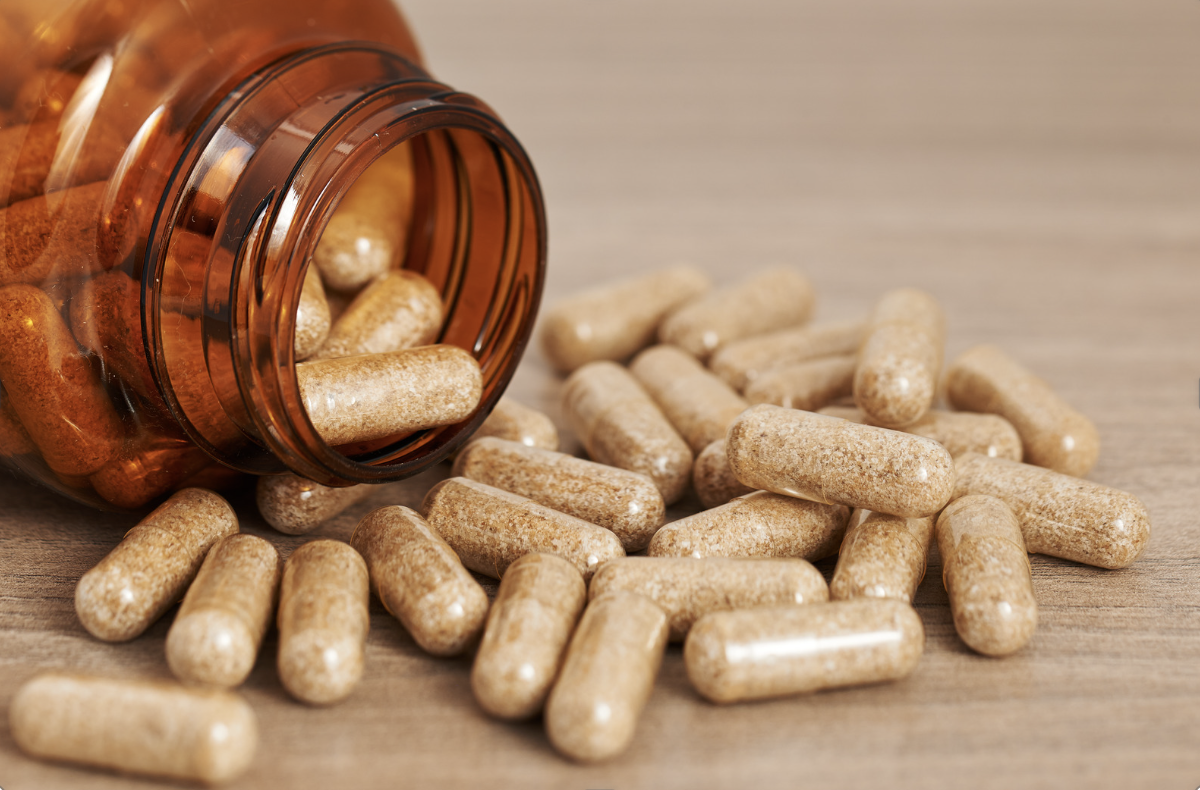 An open amber pill bottle on a wooden surface with beige capsules spilling out.