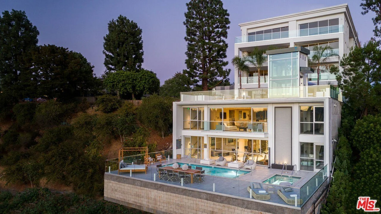 Luxury modern multi-story house with glass walls, outdoor pool, hot tub, and outdoor seating area, situated on a hillside surrounded by trees at twilight.