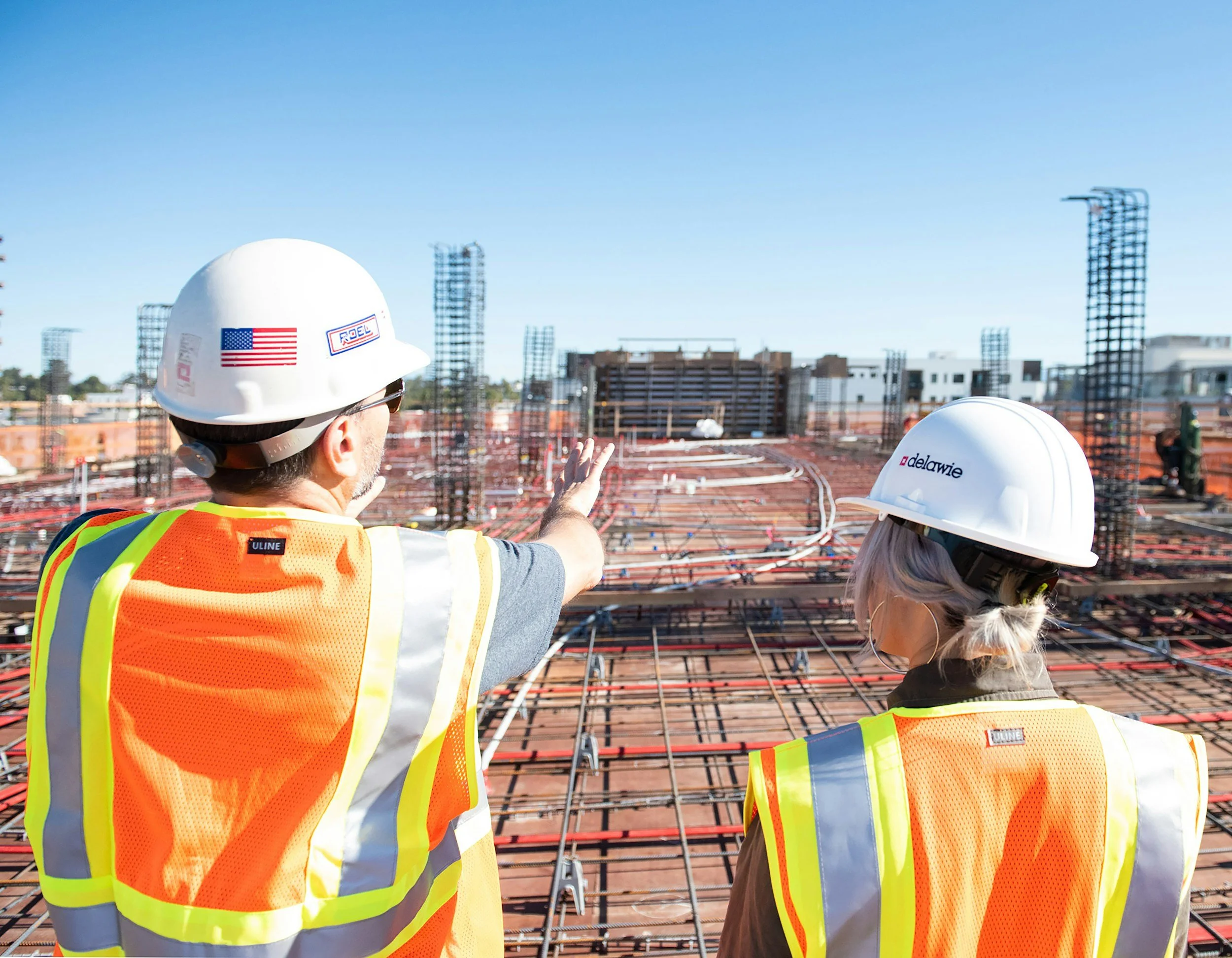 Two construction workers in safety vests and helmets inspecting a construction site with steel rebar. One worker is pointing towards the site. In the background, there are buildings and a clear blue sky.