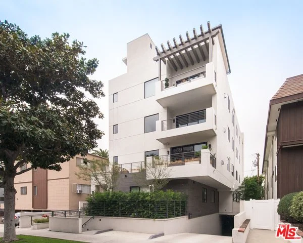 Modern multi-story white apartment building with balconies and rooftop pergola, surrounded by trees and neighboring structures.