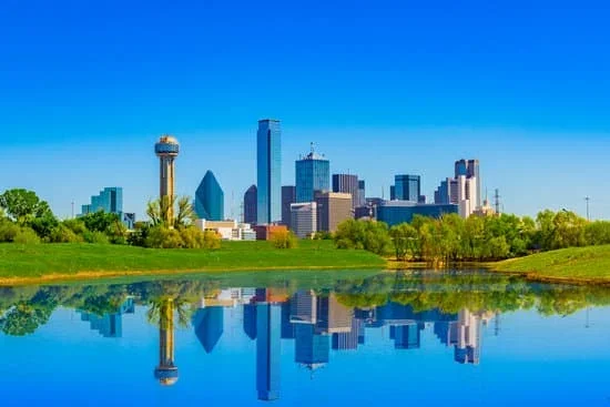 Dallas skyline with tall buildings and a water reflection, green trees and grass in the foreground, clear blue sky.