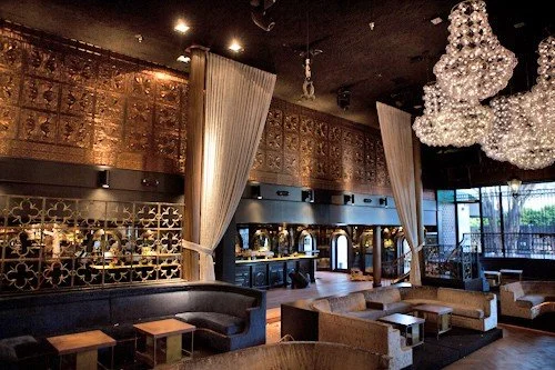 Upscale lounge with plush seating, gold decor, white curtains, and a large chandelier.