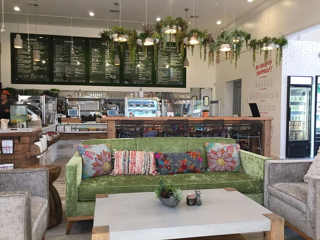Interior of a cozy cafe with a green vintage velvet sofa decorated with floral and striped cushions, a white marble coffee table with a small plant arrangement, and a high counter with barstools. Behind the counter, there is a large black menu board, various coffee and food equipment, and hanging planters with greenery. The wall features a red sign that reads "Be good to yourself!" and a glass-door refrigerator on the right side.