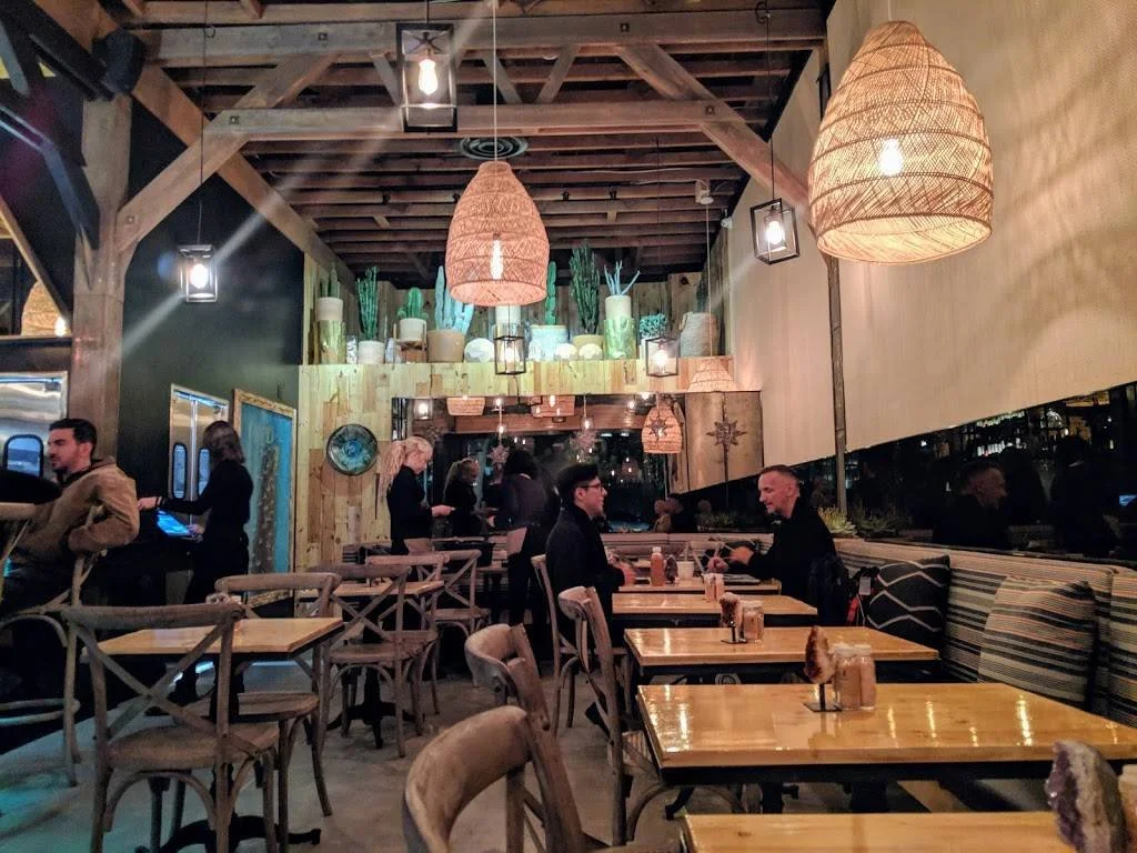 Interior of a cozy restaurant with wooden furniture, large woven pendant lights, potted cacti, and patrons dining and waiting in line.