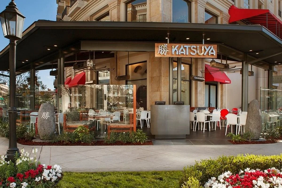 Exterior of a restaurant named Katsuya with outdoor seating, glass walls, and red umbrellas. There are flowers and shrubs in front and a black streetlamp.