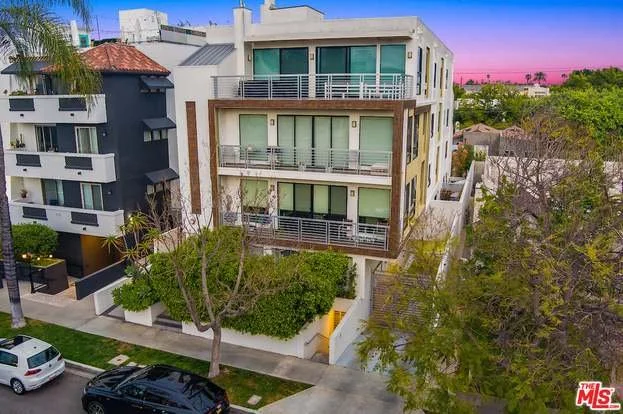 Modern multi-story residential building with balconies, surrounded by trees and parked cars, with a colorful sunset sky in the background.