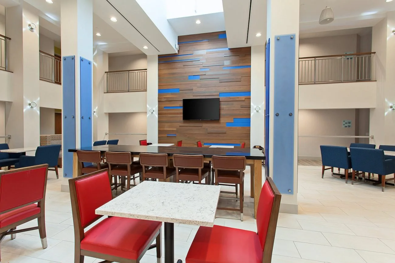 Interior view of a modern hotel lobby or lounge area featuring seating, a wall-mounted TV, wood paneling with blue accents, and multiple levels with balconies.