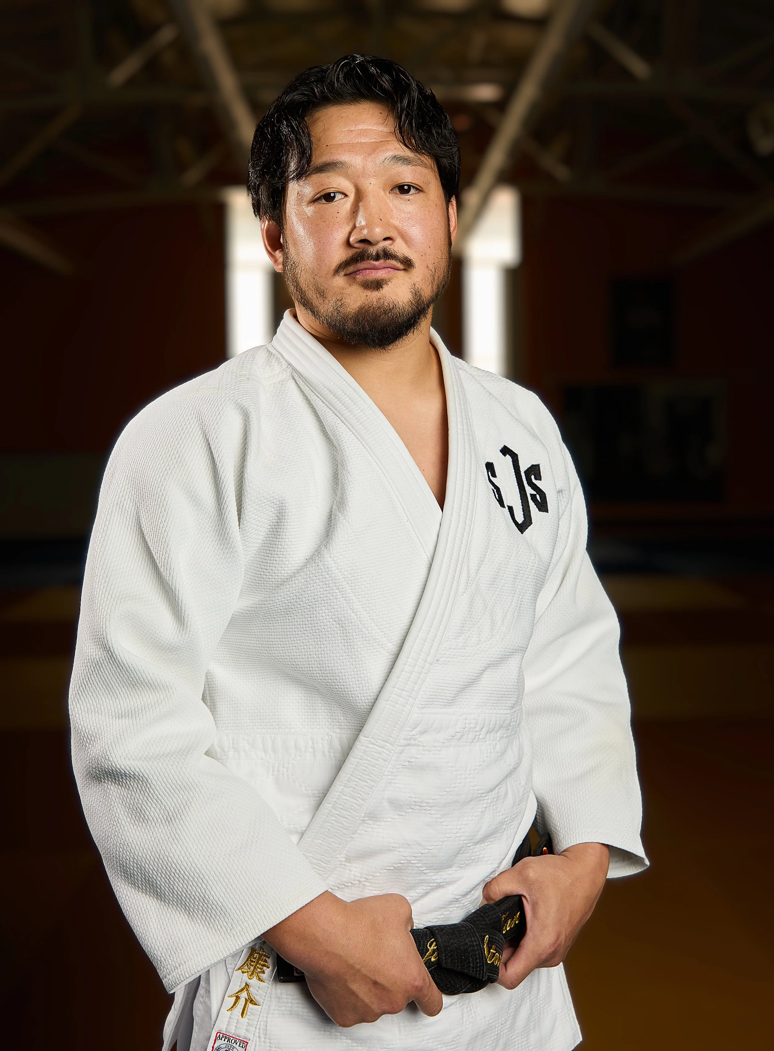 A man wearing a white judo gi with black belts stands against a black background, smiling at the camera.