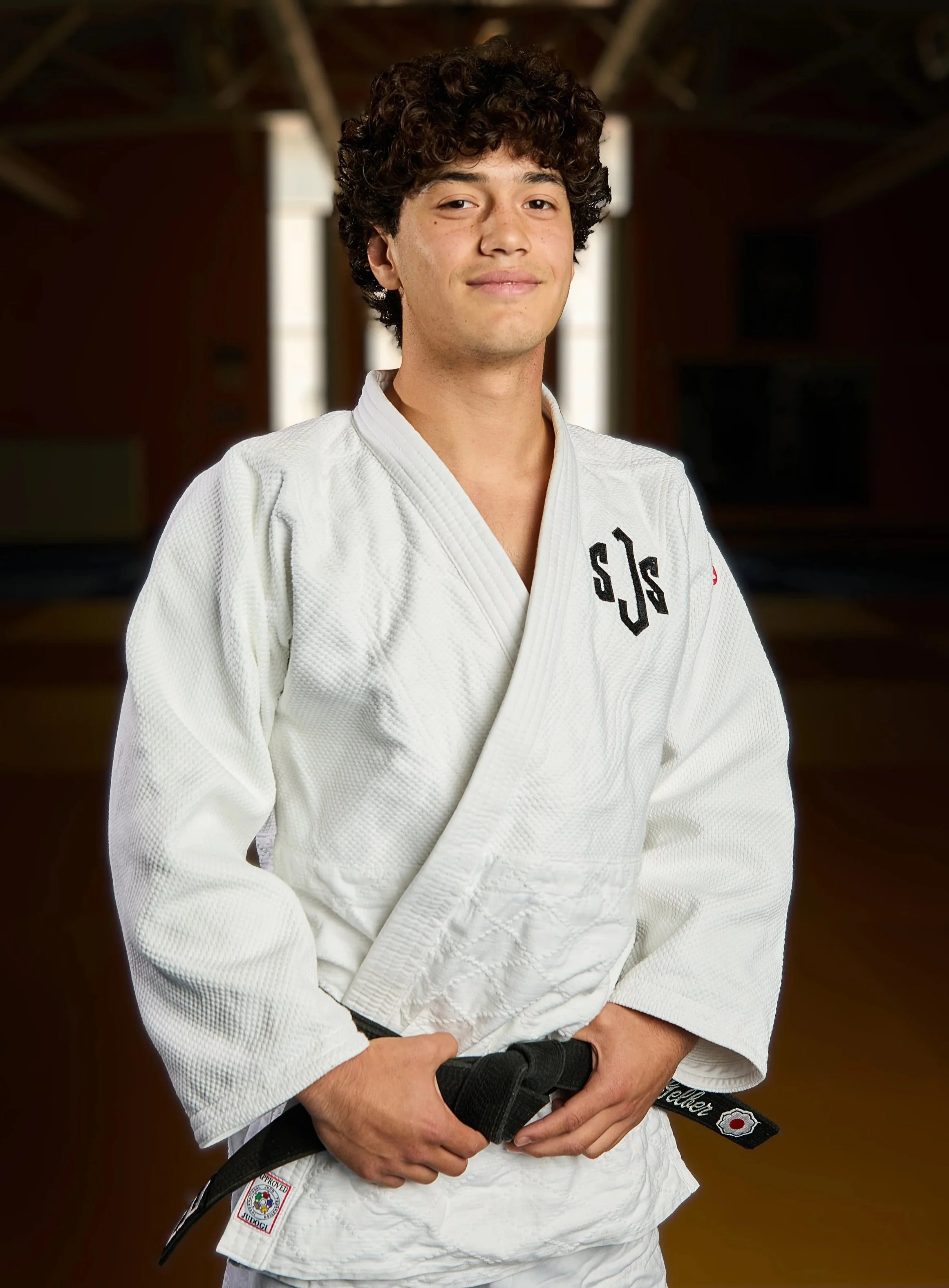 A young man wearing a white judo gi and a brown belt, standing against a dark background, holding his belt with both hands.