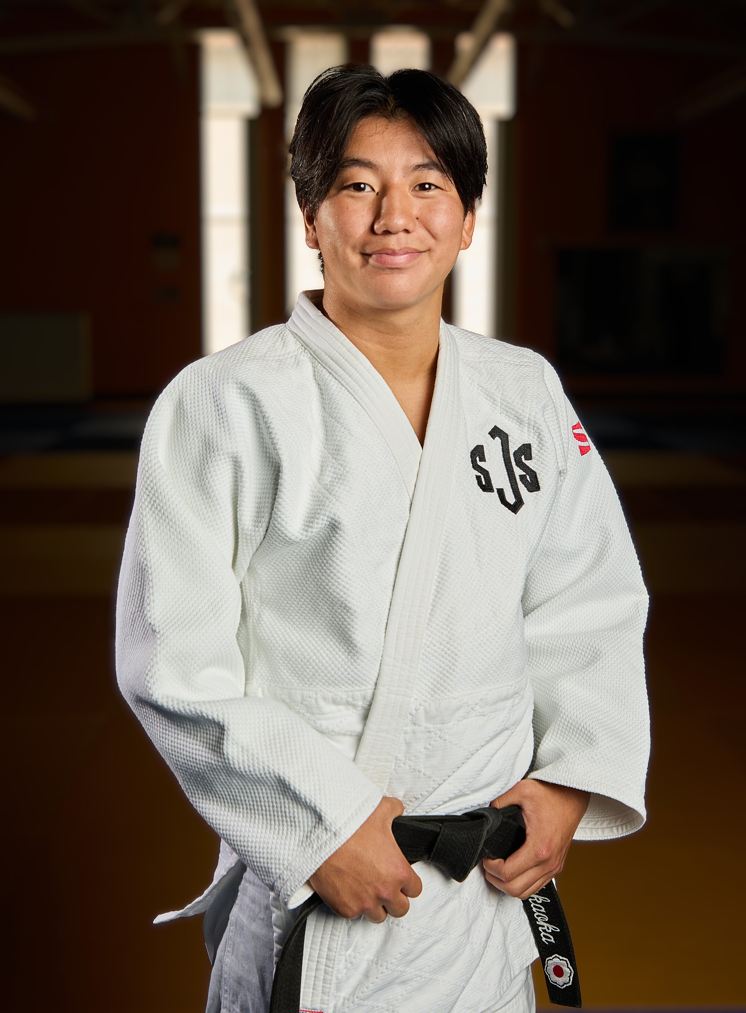 A young woman in a white Jiu-Jitsu gi with a black belt, smiling at the camera against a dark background.
