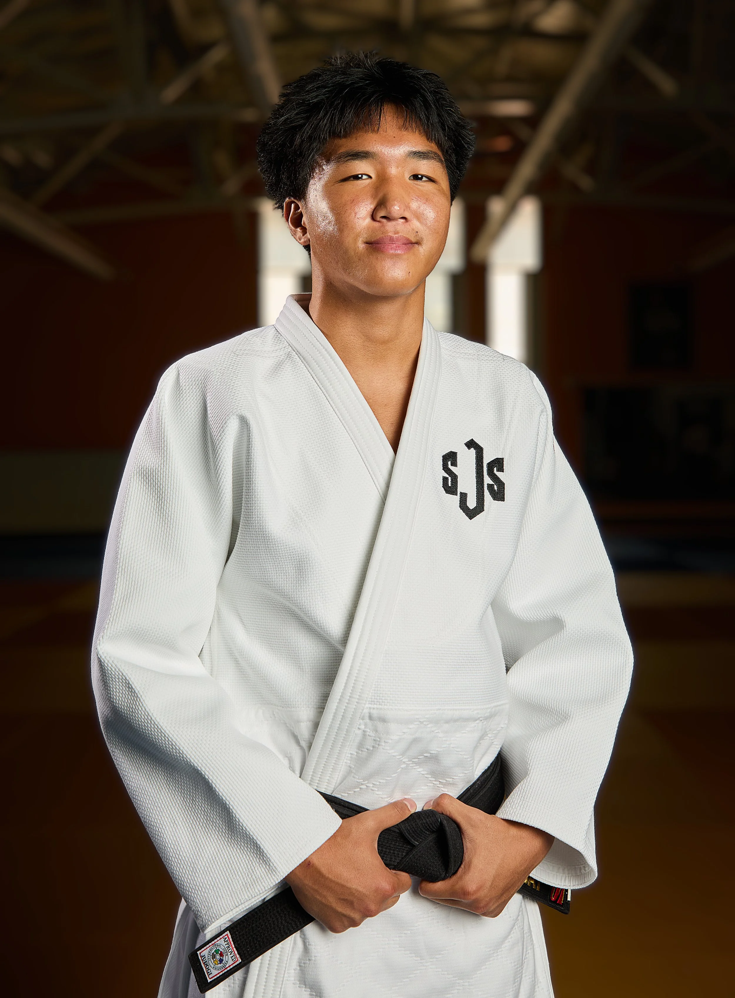 A young man dressed in a white Brazilian Jiu-Jitsu gi, standing against a black background, smiling and holding his black belt.