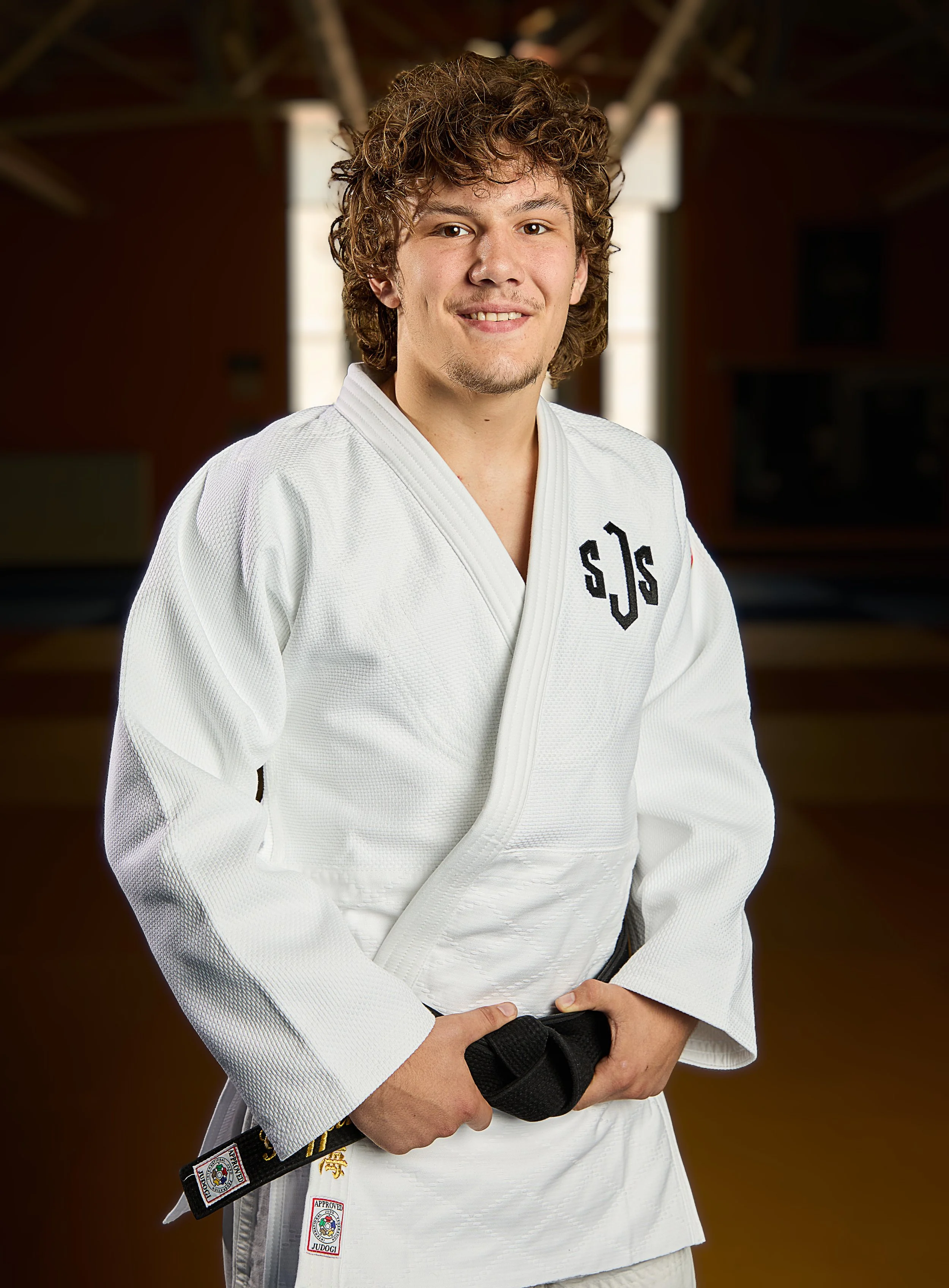 Young male martial artist in a white gi with a black belt, standing against a black background, looking determined.