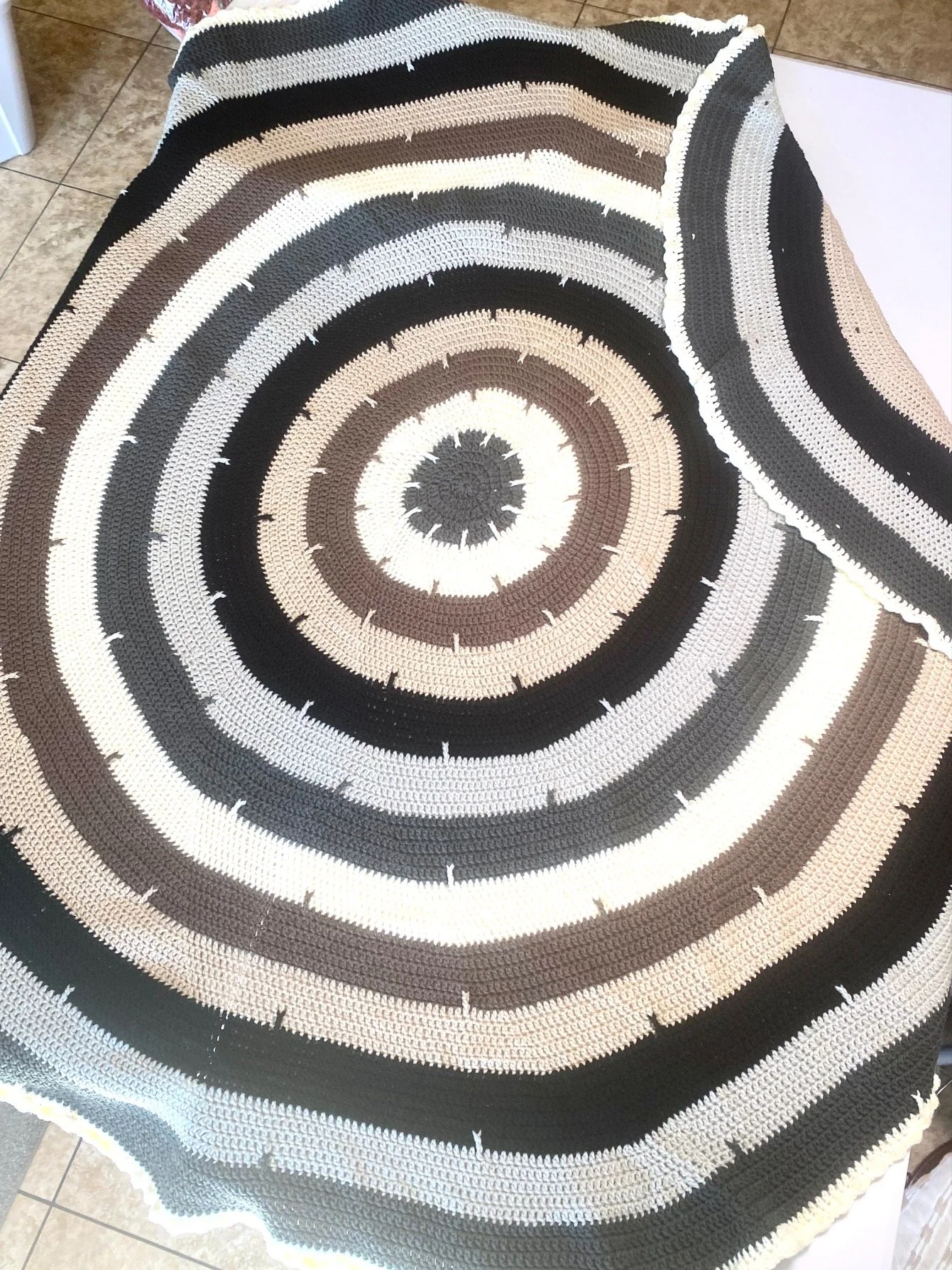 A circular crochet blanket with concentric rings in black, white, gray, and brown, laid flat on a surface.