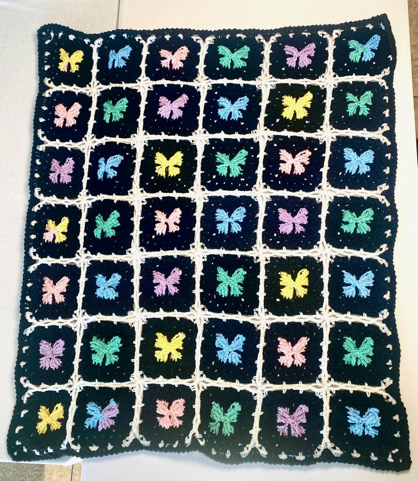 Crocheted blanket with pastel-colored butterfly embroidery in a grid pattern on black background.