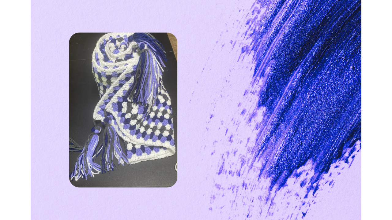 A close-up image of a crocheted scarf with blue, white, and gray colors and fringed ends, placed on a black surface with a purple paint streaked background.