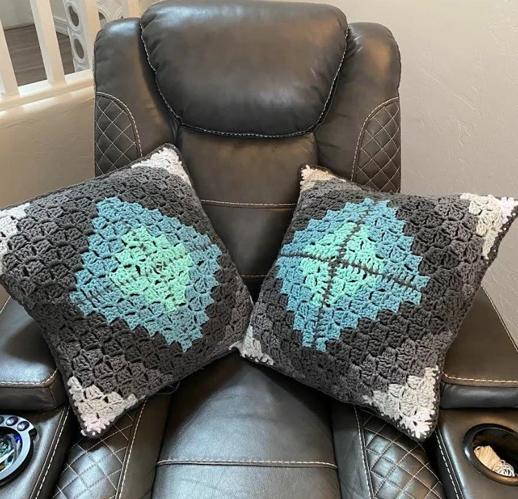 Two crocheted throw pillows with a heart design in shades of blue, green, and purple, placed on a black leather recliner.