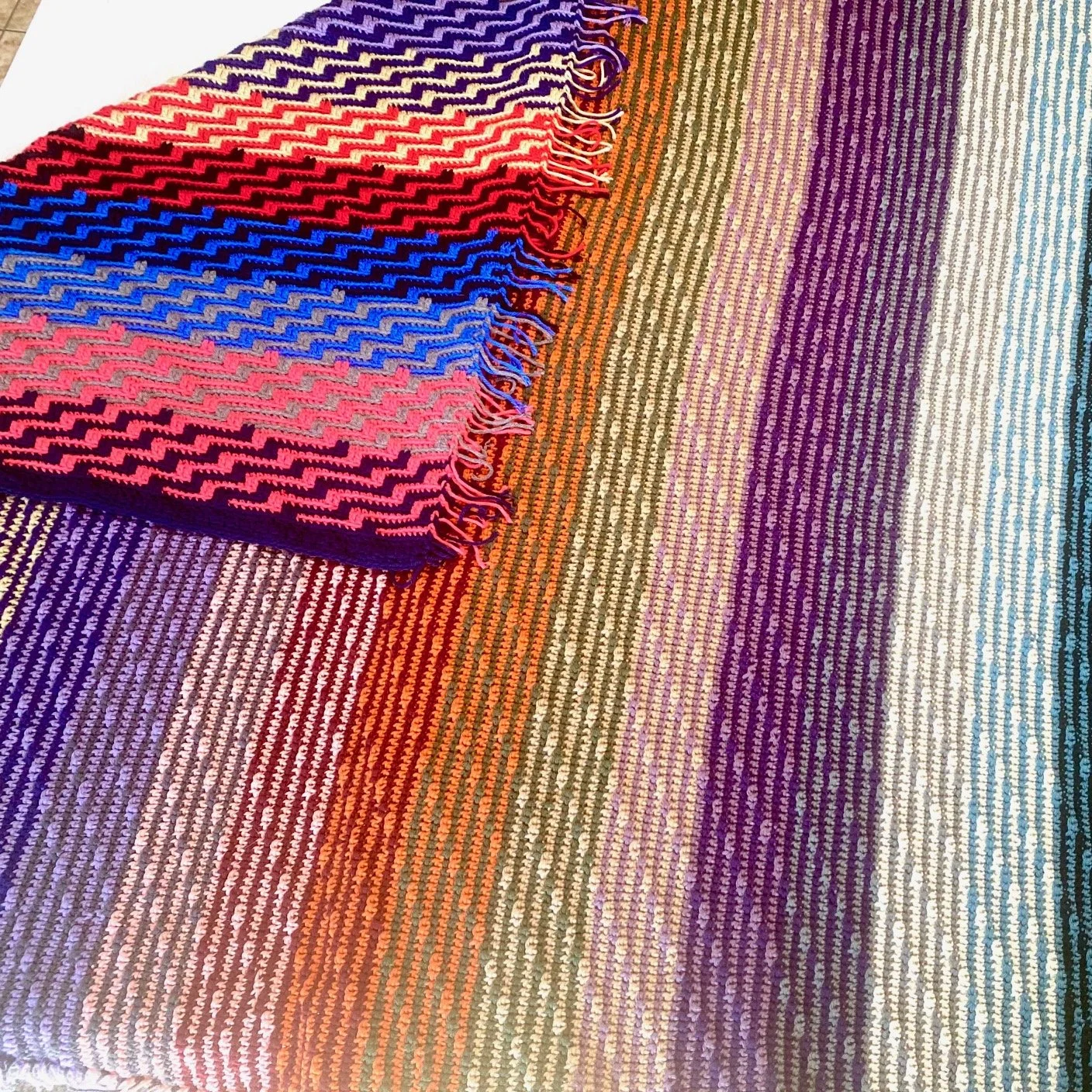 A colorful woven blanket with stripes of purple, pink, red, orange, yellow, green, blue, and white. A folded section of the blanket with a zigzag pattern and fringes is placed on top of the striped blanket.