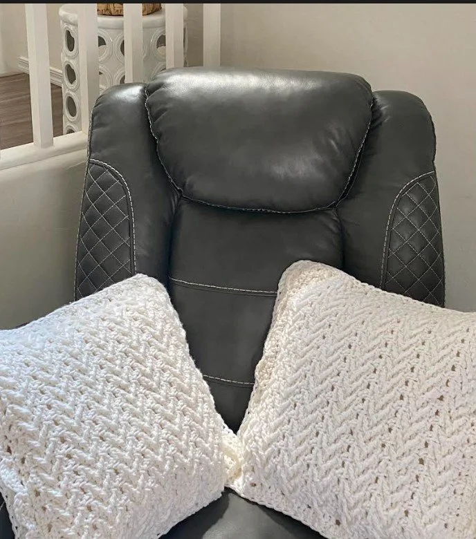 A black leather recliner chair with two white knitted pillows on it, placed in a living room near a white railing.