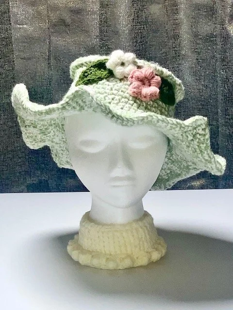 A white mannequin head wearing a crocheted hat with a wide, floppy brim decorated with pink and white flowers and green leaves, set against a dark wooden background.