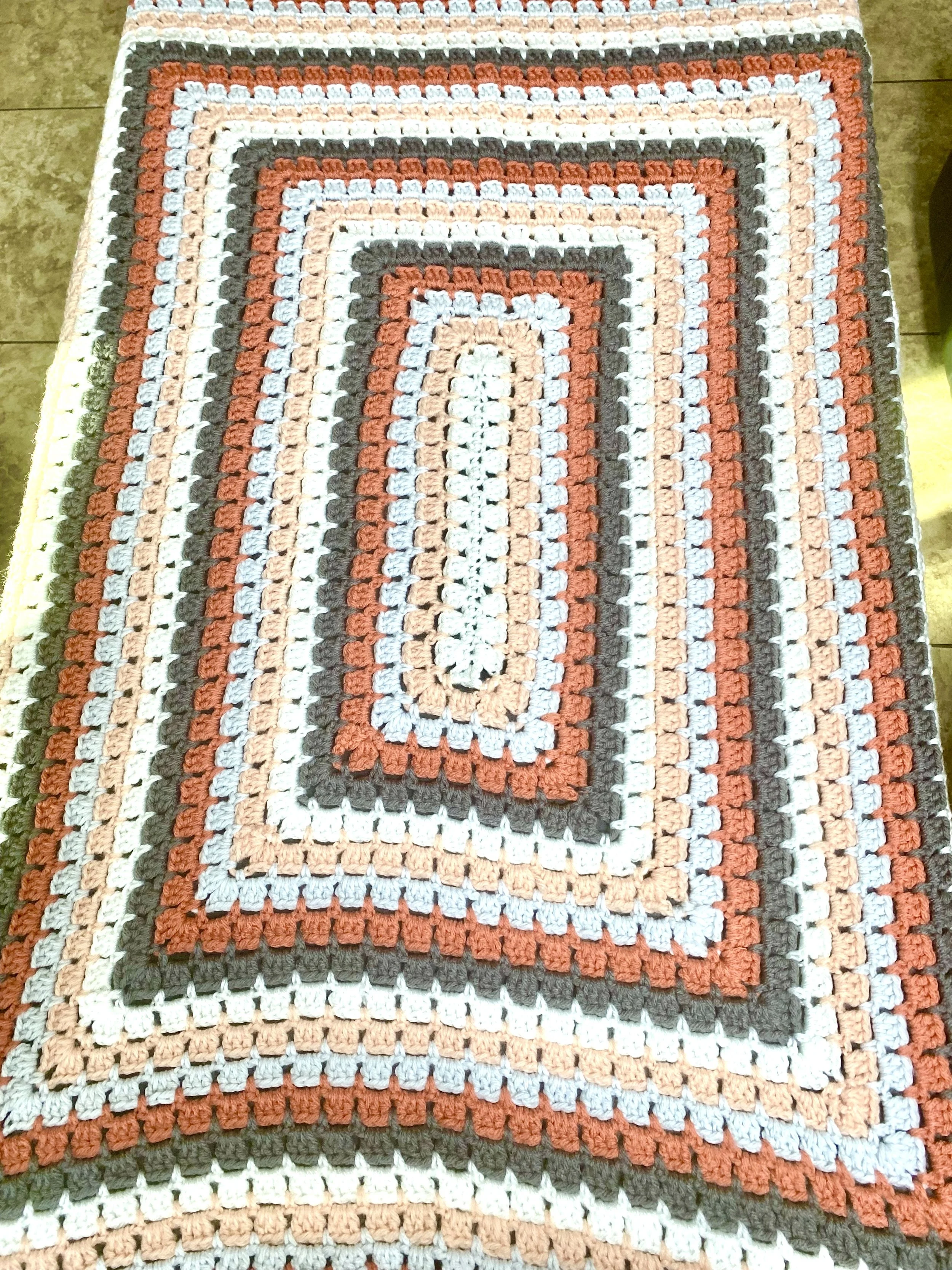 A crochet rug with geometric striped pattern in shades of white, beige, brown, black, and orange, placed on a tiled floor.